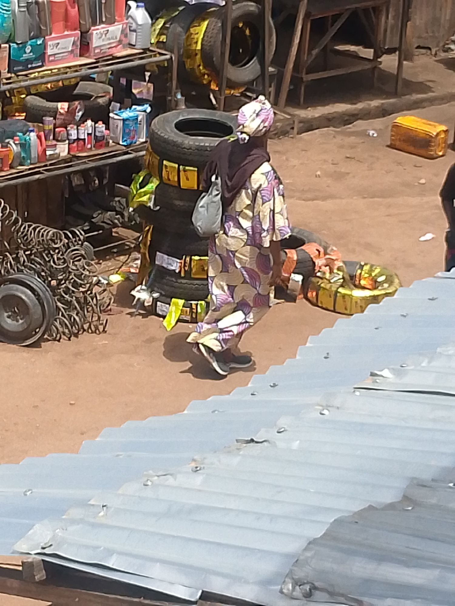 The image shows an outdoor scene in Bukuru, Nigeria. A woman wearing a patterned headscarf and clothing, along with a shoulder bag, walks past a roadside automotive shop. The shop displays tires stacked and propped up and automobile parts on shelves. There are various liquids and containers on the shelves. Used tires, a pile of coiled metal springs, and other spare parts are on the ground. A yellow plastic container sits nearby. In the foreground is a corrugated metal roof.