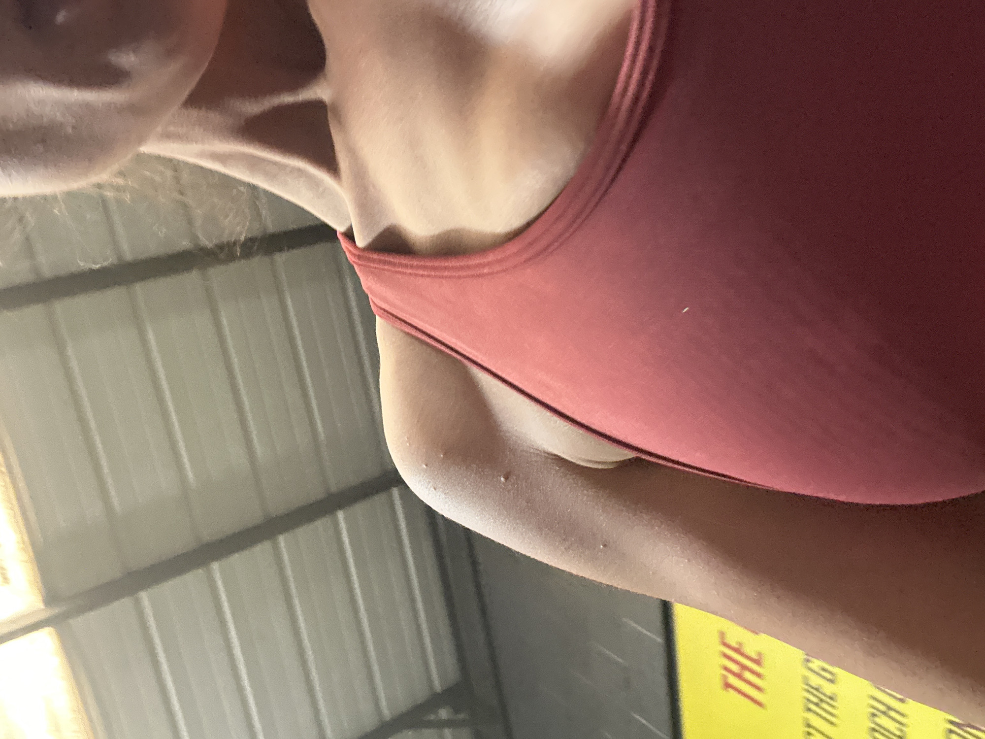 The image shows a close-up view of a woman's upper body, specifically her neck, shoulder, and part of her arm. She is wearing a red tank top. The skin is light-toned and shows minor bumps or imperfections. The background features a corrugated metal ceiling or wall, with a blurred yellow sign visible at the bottom. The location is not discernible from the image and cannot be determined to be Tonbridge and Malling, United Kingdom.