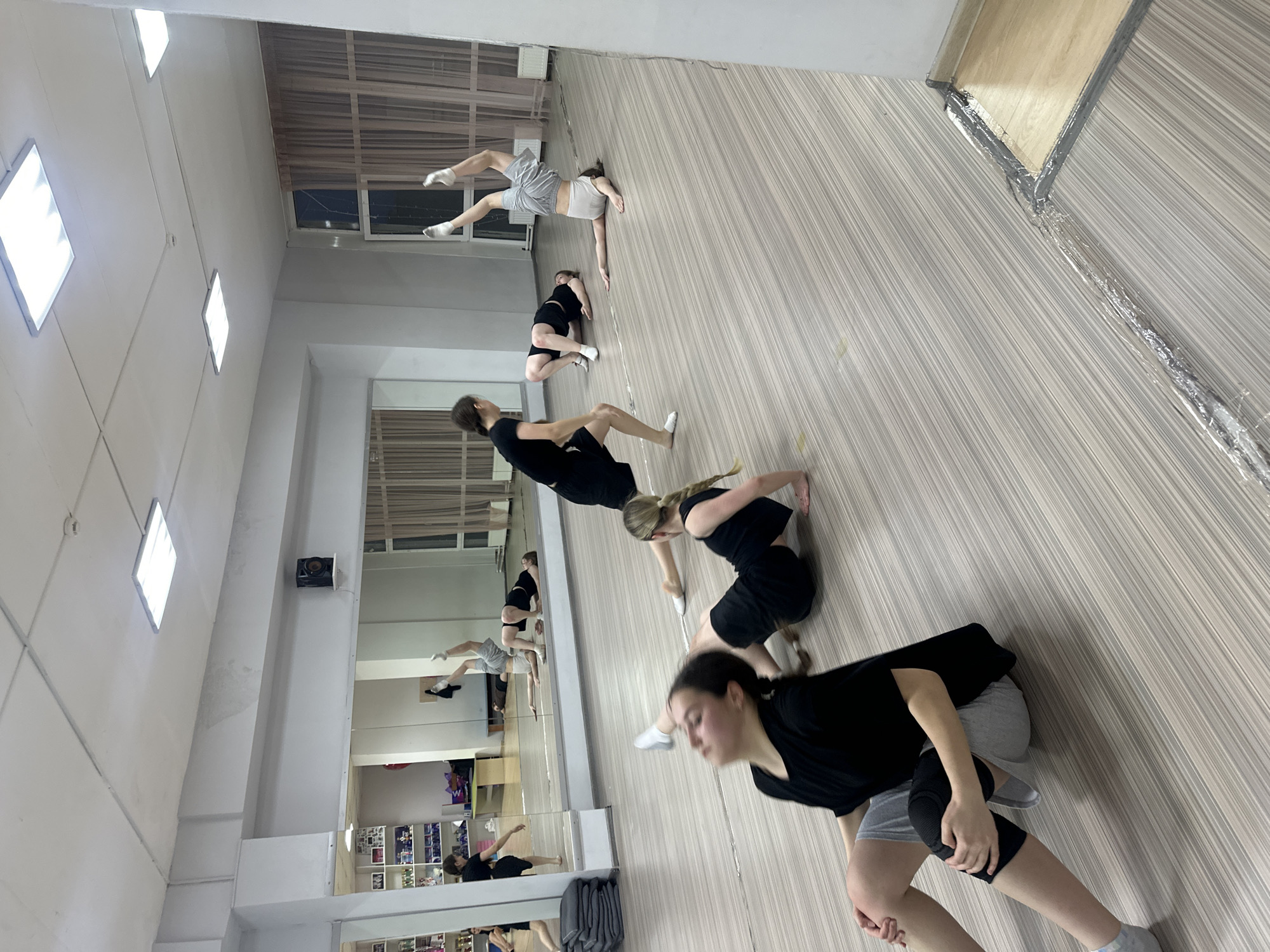 The image captures an indoor dance or fitness studio in Votkinsk, Russia, during what appears to be a practice session. The room features a light-colored wooden or laminate floor with a distinct striped pattern, and white walls. A prominent mirrored wall on the left side reflects the interior of the studio and the individuals present.
Several young people, predominantly females, are visible within the space, engaged in various dynamic poses on the floor. They are dressed in athletic attire, including black t-shirts, black and grey shorts, sports bras, and socks; some also wear black knee pads. Their stances suggest stretching, warm-up exercises, or specific dance movements. In the foreground, one individual in a black top and grey shorts with knee pads is in a crouching position, appearing focused. Further back, another person in a light grey sports bra and grey shorts is captured in a more elevated, dynamic stretch. Reflections of additional participants and studio items, such as shelves with belongings, are visible in the mirrors.
The studio is brightly lit by several rectangular fluorescent light fixtures embedded in the white ceiling. High windows are visible on the back wall, suggesting an external view or another room, but no outdoor elements are clearly discernible. Visible along the upper walls are some utility conduits or ventilation components. The general mood is one of focused activity and discipline. No visible text is present in the image.