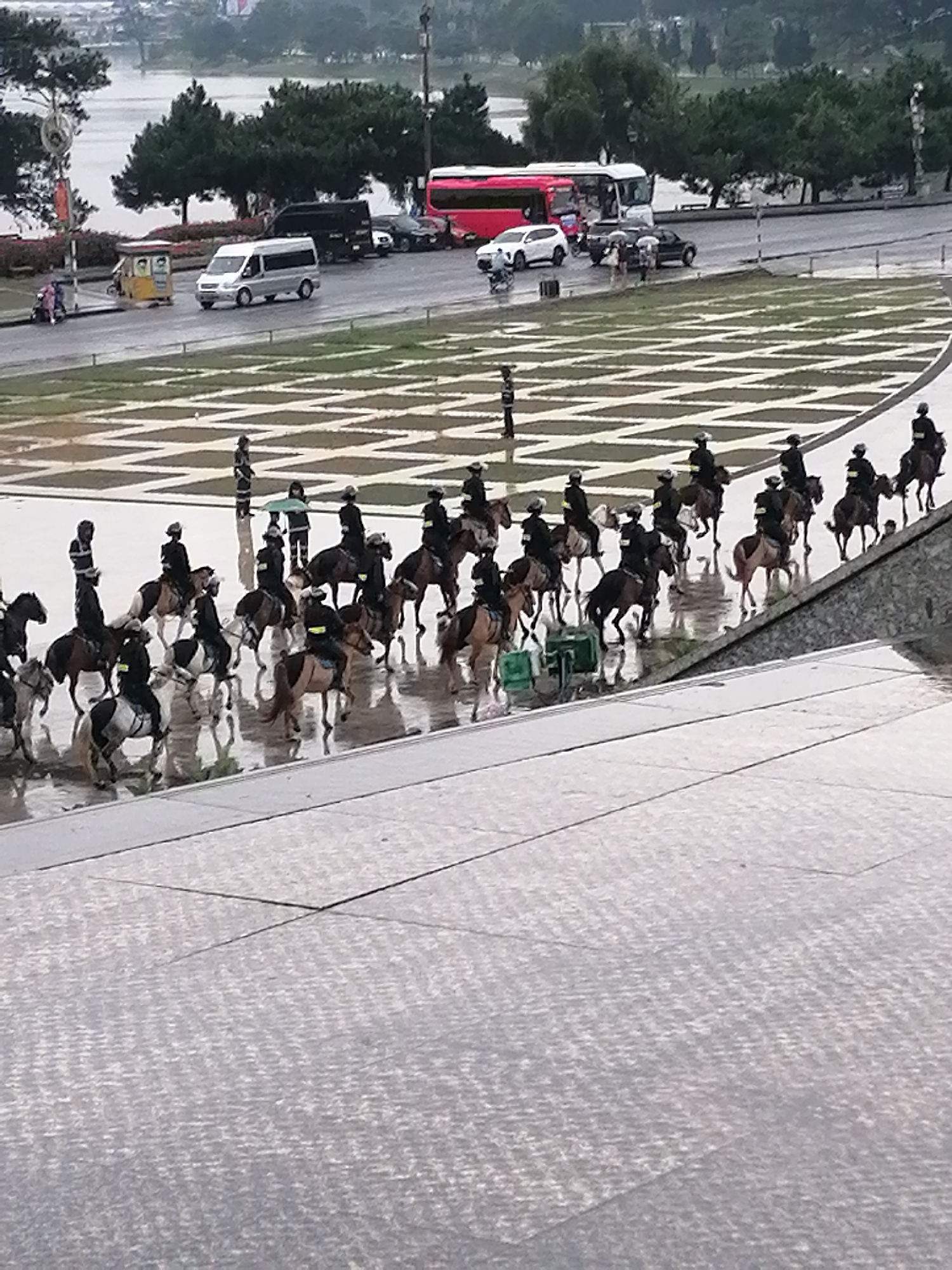 The image depicts a squad of mounted police officers on horseback, positioned on a wet, reflective surface, likely a paved plaza or thoroughfare. The officers are all wearing dark uniforms and helmets, and are riding dark-colored horses. They appear to be lined up in formation, suggesting a ceremonial or security detail. The environment appears to be overcast, with the wet surfaces indicating recent rain or damp conditions. In the background, several vehicles are visible, including a silver van, a black SUV, and a red bus, suggesting an urban or semi-urban setting. Some trees and distant buildings are also discernible. The scene is likely in Da Lat, Vietnam, given the context provided. The overall impression is one of order and preparedness.