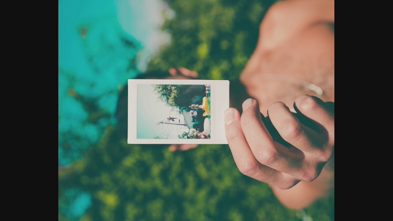 A close-up shot shows a person's hand holding up a Polaroid photograph in front of their face. The photograph depicts a scene of people in a backyard, with one person jumping off a ledge into a pool, while others are seen near inflatable pool toys. The background of the main image is blurred green foliage, with a vibrant teal sky. The person's torso, which is bare, is visible behind the photograph, and they are wearing a silver chain.