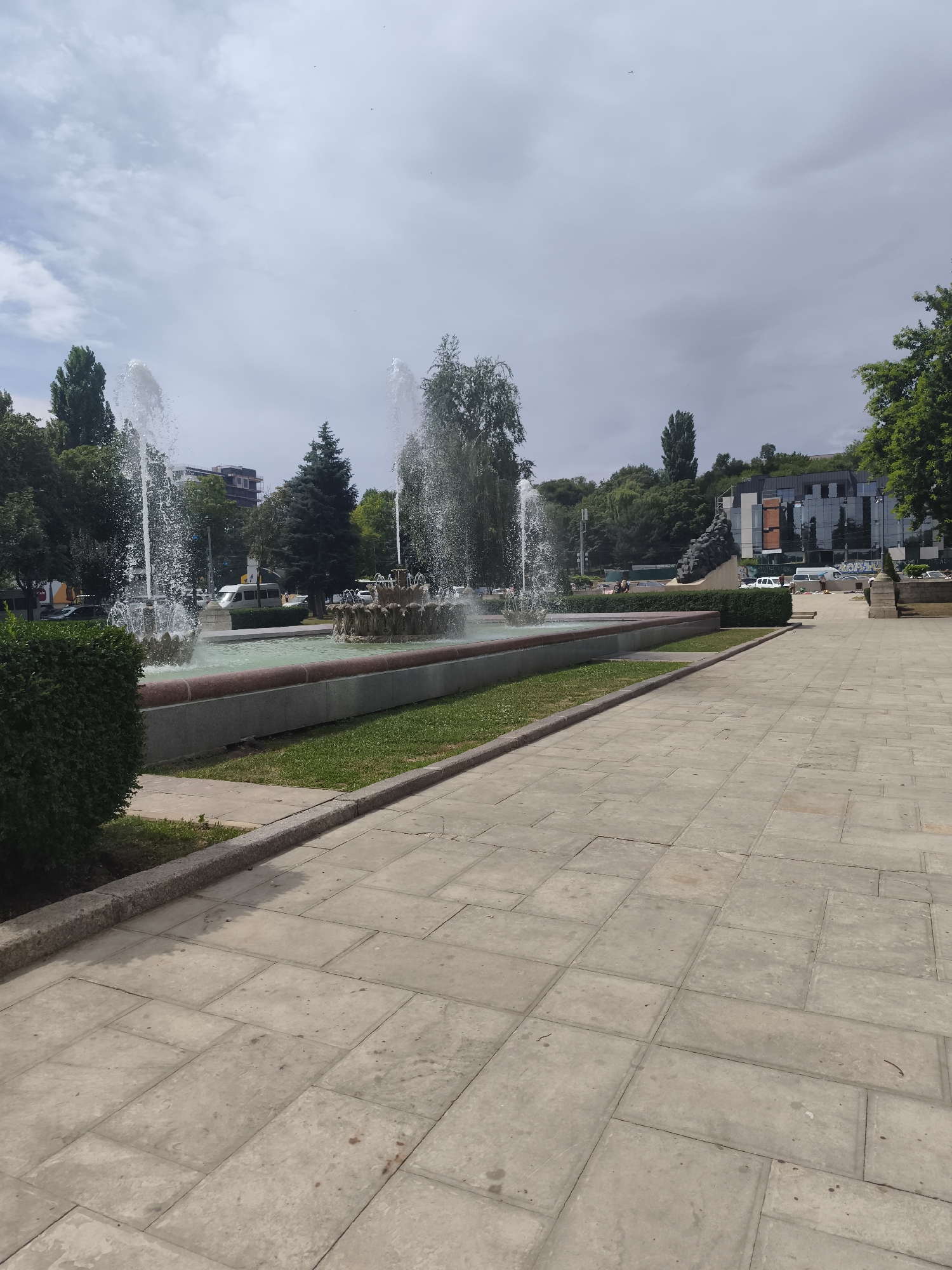 The image shows a wide, paved plaza in front of a large fountain with multiple jets of water spraying upwards. The fountain has a long, rectangular basin with a stone rim. Behind the fountain, there are green trees and some buildings, including a modern building with a glass facade. A few vehicles, including a white van, are visible on a street in the background. The sky is overcast with scattered clouds. The plaza is made of large, light-colored square and rectangular paving stones. A manicured hedge is visible on the left side of the plaza. The scene appears to be a public space, possibly a park or a square in a city. This is a view of the Stephen the Great Central Park fountain in Chișinău, Moldova.