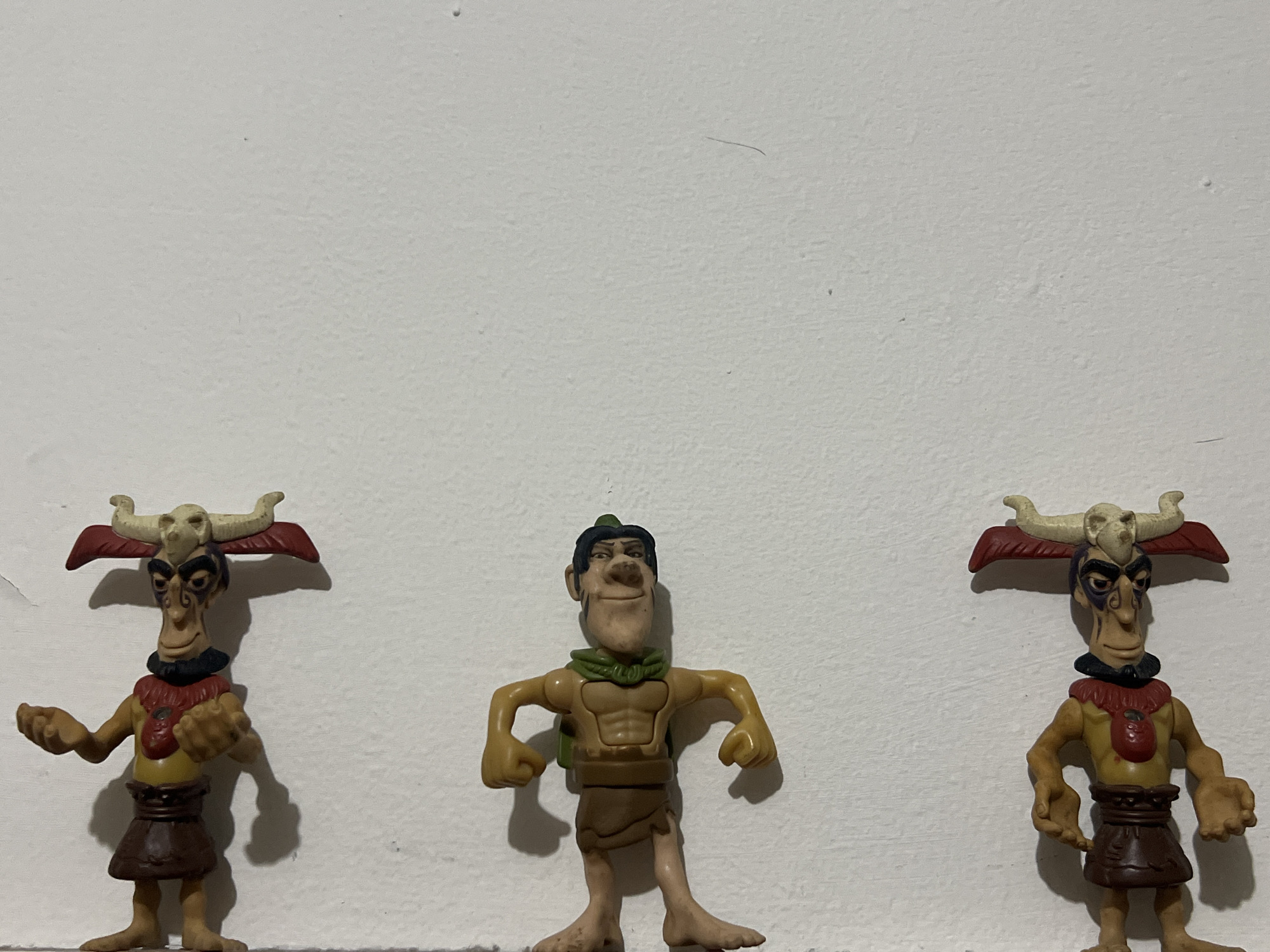 The image features three plastic toy figures arranged in a row against a plain, light-colored, textured wall. The central figure is a male character with dark hair, a green scarf, a muscular bare torso, and a brown loincloth. He stands with his arms slightly bent and fists clenched.
On either side of the central figure are two identical tribal or shaman-like characters. These figures have elongated faces with black and purple markings around their eyes, a black necklace, a red textured collar, and a brown skirt-like garment. Each is adorned with a skull and horns on its head, accompanied by red, wing-like decorative elements. Both flanking figures stand with their hands open, palms facing upwards.
The figures are stationary, displayed against the wall, which exhibits a slightly bumpy texture and a few minor imperfections such as small dark specks and a thin dark line in the upper portion. Shadows are cast behind the figures, suggesting a light source from the front. The scene appears to be indoors, likely under artificial lighting. No specific activity or event is taking place beyond the static arrangement of the toys. No text is visible in the image. The location is unknown.