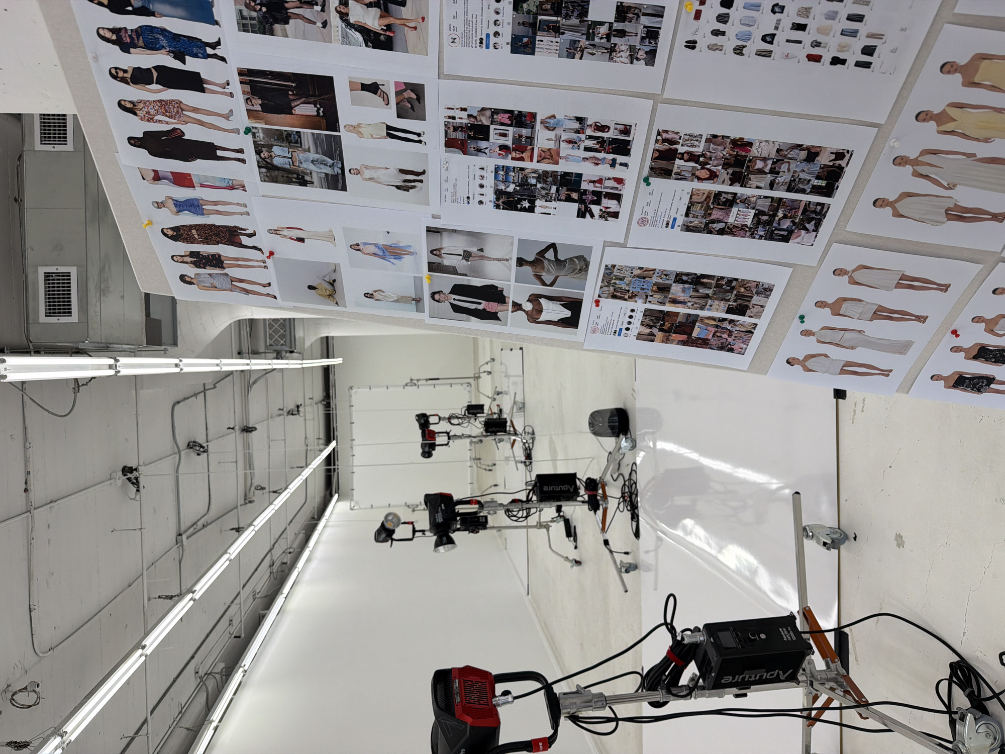 The image shows a photography studio in Los Angeles. Sheets of fashion layouts and mood boards are pinned to a wall. Lighting equipment includes Aputure brand units on stands. The studio has a high ceiling with fluorescent tube lighting. The floor is white.