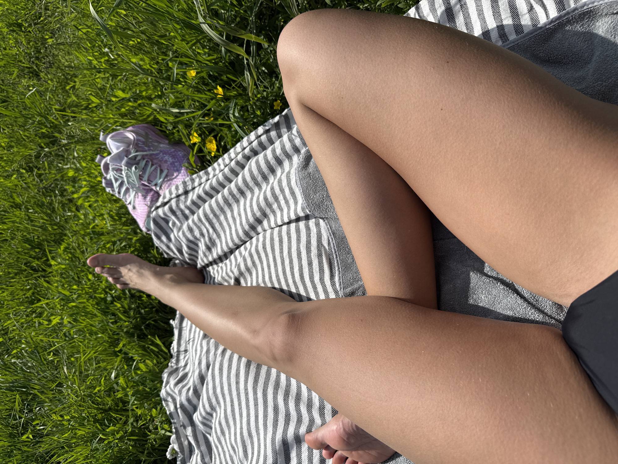 The image captures a first-person, high-angle view of a person relaxing outdoors on a sunny day in Saint Petersburg, Russia. The primary subjects are the person's legs, which are tanned and visible from the upper thighs down to the bare feet. The person is lying or sitting on a grey and white striped towel or blanket, which covers part of a vibrant green grassy area.
The setting is a natural outdoor environment, likely a park or field, characterized by lush green grass with scattered small yellow flowers, possibly buttercups, indicating springtime or early summer. Bright sunlight bathes the scene, creating distinct shadows and highlighting the texture of the grass and the person's skin.
One notable detail is the presence of piloerection (goosebumps) on the person's skin, particularly visible on the knees and thighs, suggesting a slight chill or perhaps a recent change in temperature despite the sunny conditions. A pair of light purple or lilac sneakers with light blue laces is placed on the grass near the towel. The edge of a dark, possibly black, swimsuit bottom is visible on the right side of the frame, near the upper thigh, implying the person might be sunbathing or dressed for warm weather activities. The overall mood conveyed is one of leisure and relaxation in nature. There is no visible text in the image.