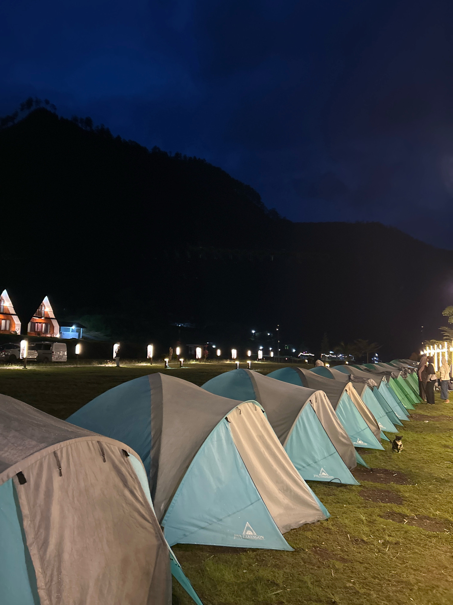 The image depicts a campsite at dusk in Merodot, Indonesia. In the foreground, a row of blue and gray tents are set up on a grassy field. The tents appear to be of the dome style, with some showing branding. Behind the tents, a line of pole lights illuminate the pathway, and several people are visible walking and standing. Further in the background, there are several A-frame houses with lights on, suggesting accommodation. A dark, tree-covered mountain range dominates the upper portion of the image, its silhouette stark against the deep blue twilight sky. The overall scene conveys a peaceful, outdoor setting as night falls.