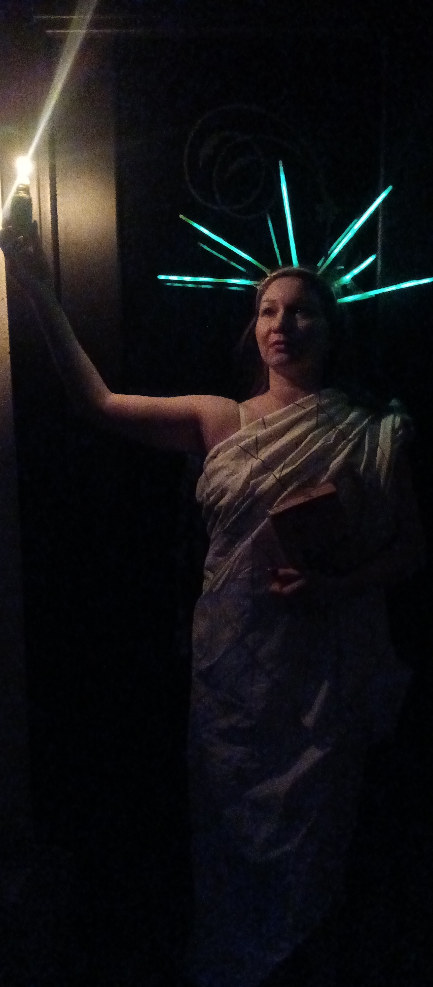 A person dressed in a costume reminiscent of the Statue of Liberty stands in a darkened environment. The person wears a draped, light-colored garment and a headdress with radiating, glowing green rods. They hold a lit object aloft in their raised right hand and a rectangular object close to their body in their left hand. The background is dark, but a textured, swirling design is faintly visible above and to the right of the person's head, while a wall is visible to their left. The location is Unknown, Unknown, and no city is visible.