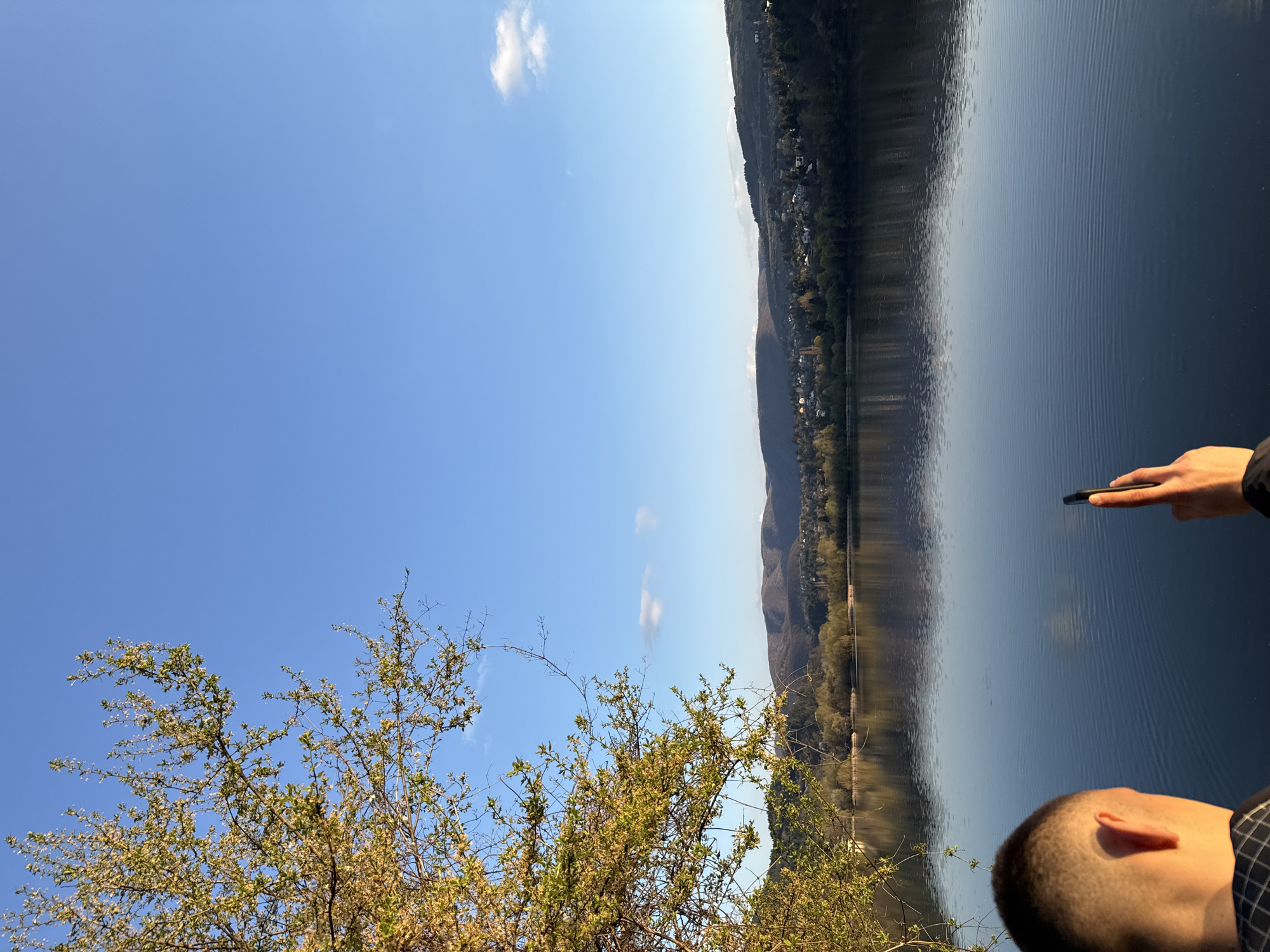 The image captures a scene in Pancharevo, Bulgaria, featuring a body of water reflecting the sky and distant mountains. The landscape is dominated by the lake's surface, with a forested shoreline and visible houses along the mountain slopes. In the foreground, branches of a tree with sparse foliage frame the view. A person's head and part of their hand holding a device are visible on the right side of the image. The sky is blue with a few clouds.