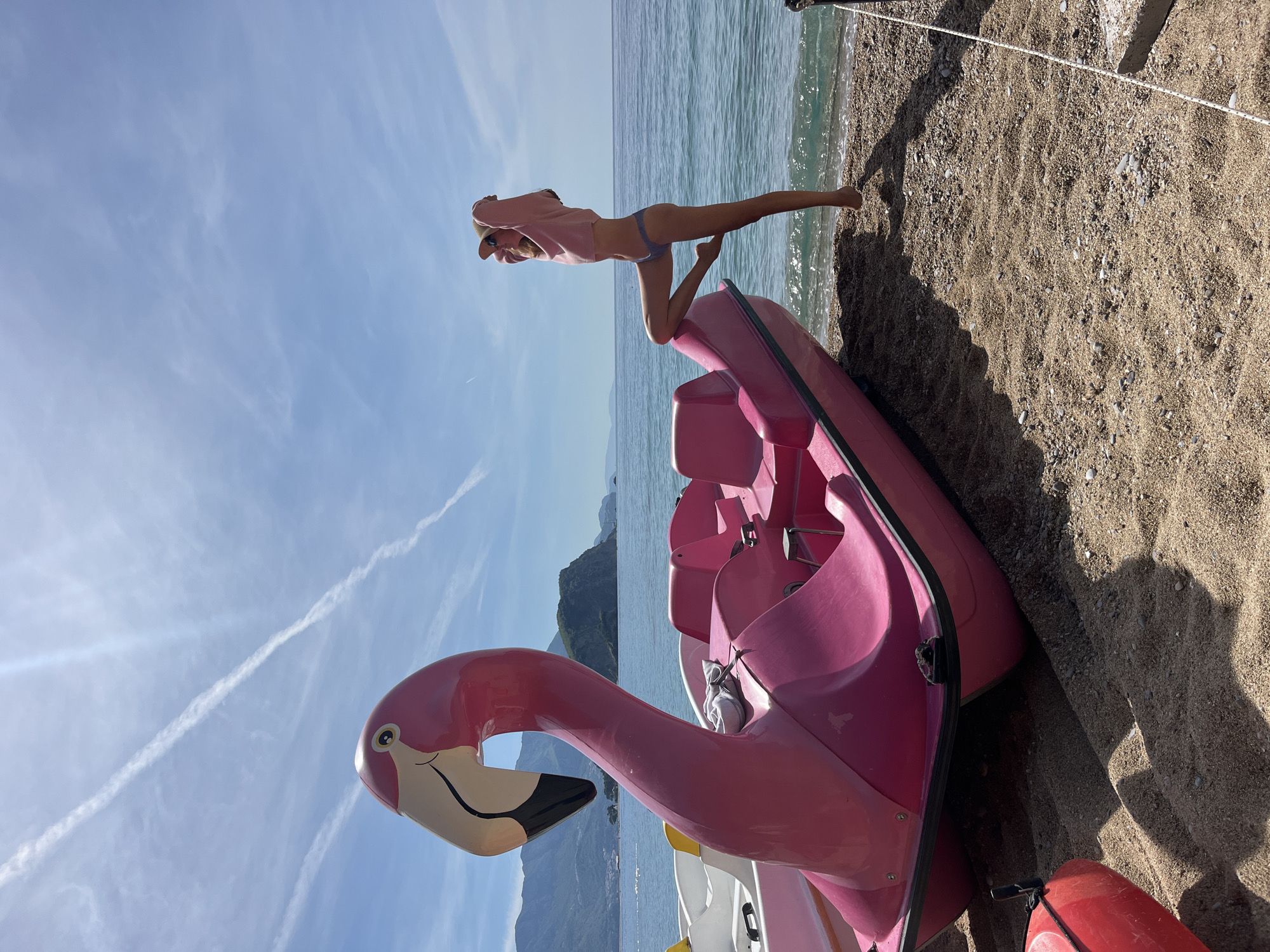 The scene is a sunny beach in Budva, Montenegro, featuring a woman striking a pose atop a pink flamingo-shaped paddle boat. The woman is wearing a pink top and blue bikini bottoms, with her body extended horizontally, supported by the boat. The flamingo paddle boat is situated on the sandy beach near the shoreline, with additional white and yellow paddle boats visible behind it. In the background, the sea stretches towards the horizon, and a mountainous landscape is partially visible. The sky is blue with sparse clouds. A rope extends from the shoreline.