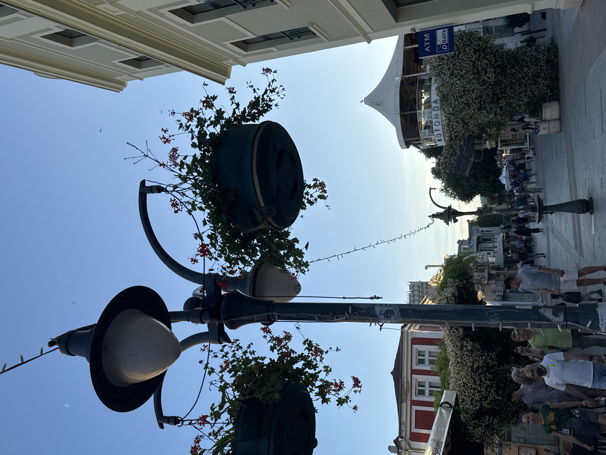 The image depicts a street scene in Burgas, Bulgaria, specifically on Bogoridi Street, known for its lively atmosphere and shops. In the foreground, a lamppost with hanging baskets of red flowers is prominent. The street itself is paved with cobblestones and lined with buildings, one of which is a cream-colored building with multiple windows. Further down the street, a white, circular structure with "ROTUNDA" signage is visible, partially covered in foliage. Several people are walking on the street, some casually strolling, while others appear to be interacting. The sky is clear and blue with a crescent moon visible. The overall impression is of a bustling urban environment on a clear day, likely late afternoon or early evening, given the lighting.