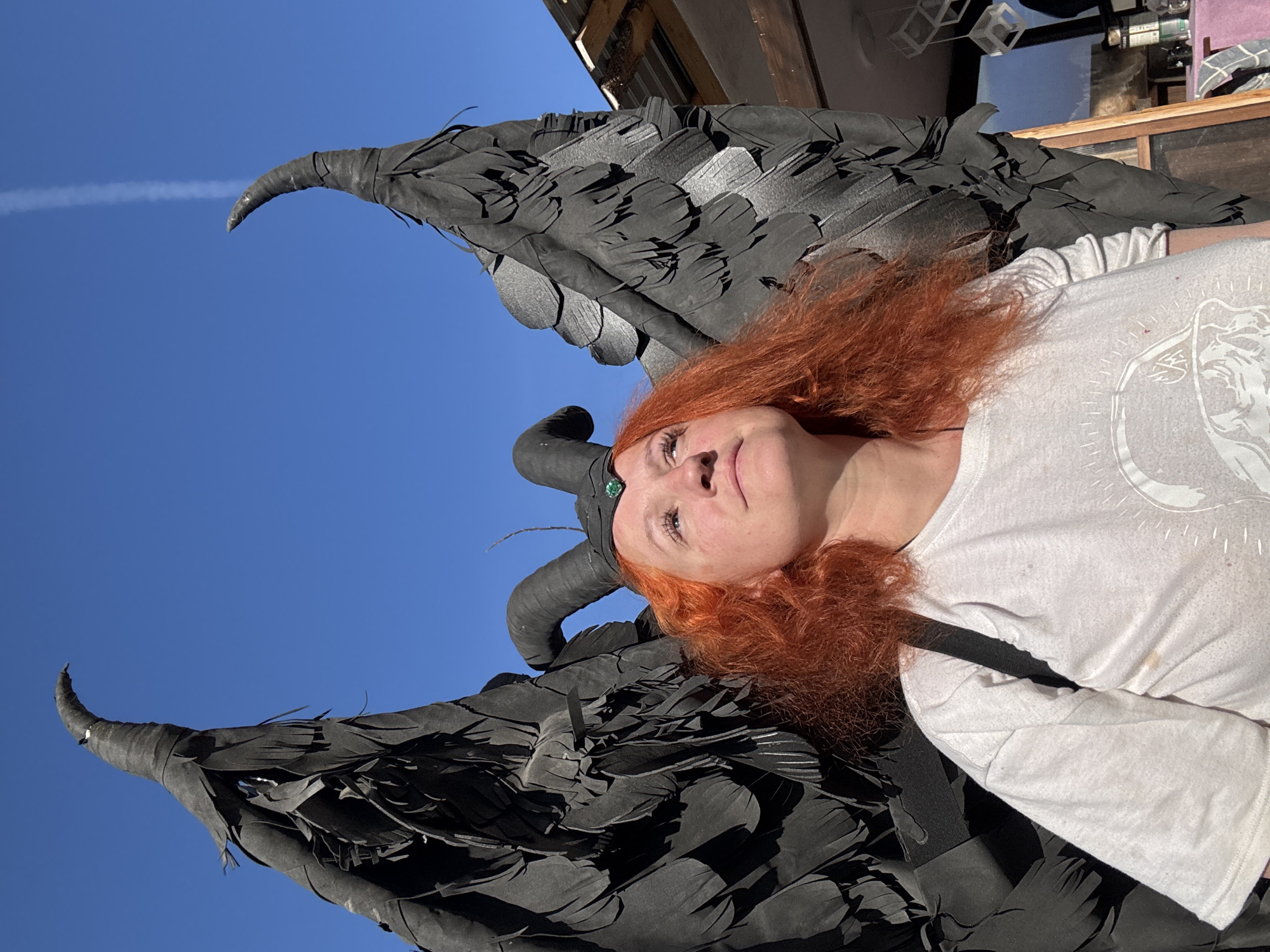 A woman with red hair and black devil horns and wings is lying down. She is wearing a white t-shirt. The sky is blue, and there is a contrail in the upper left corner. The location is not discernable from the image.