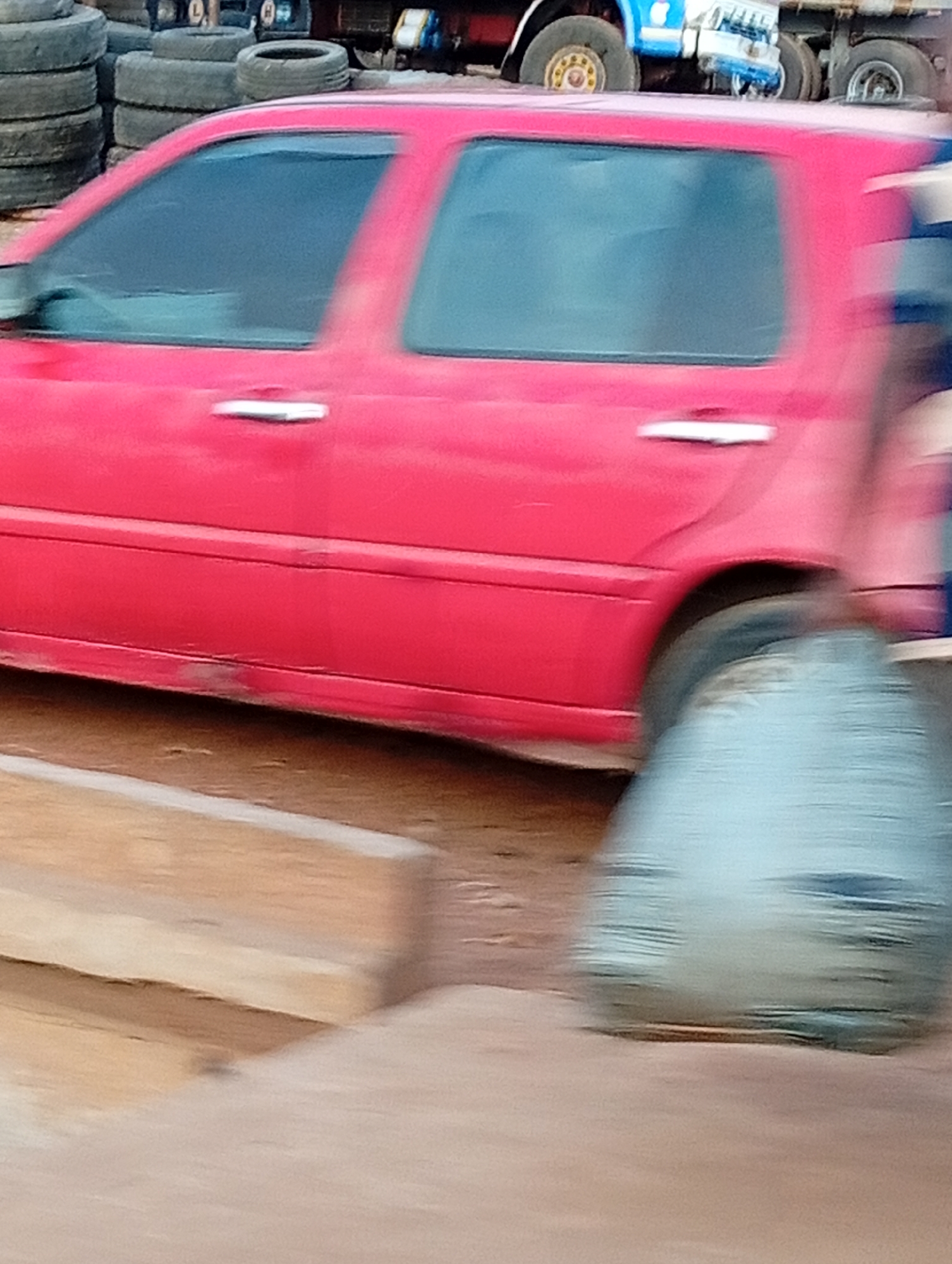 The image shows a blurry, side-on view of a red sedan-type vehicle. The location appears to be near a tire repair or disposal area, evidenced by stacks of old tires in the background. In the background, there is also a truck, possibly a flatbed, with blue and white coloring. Due to the blur, actions and interactions cannot be accurately determined, and the environment appears to be outdoors, possibly a commercial or industrial area in Nassarawa, Nigeria.