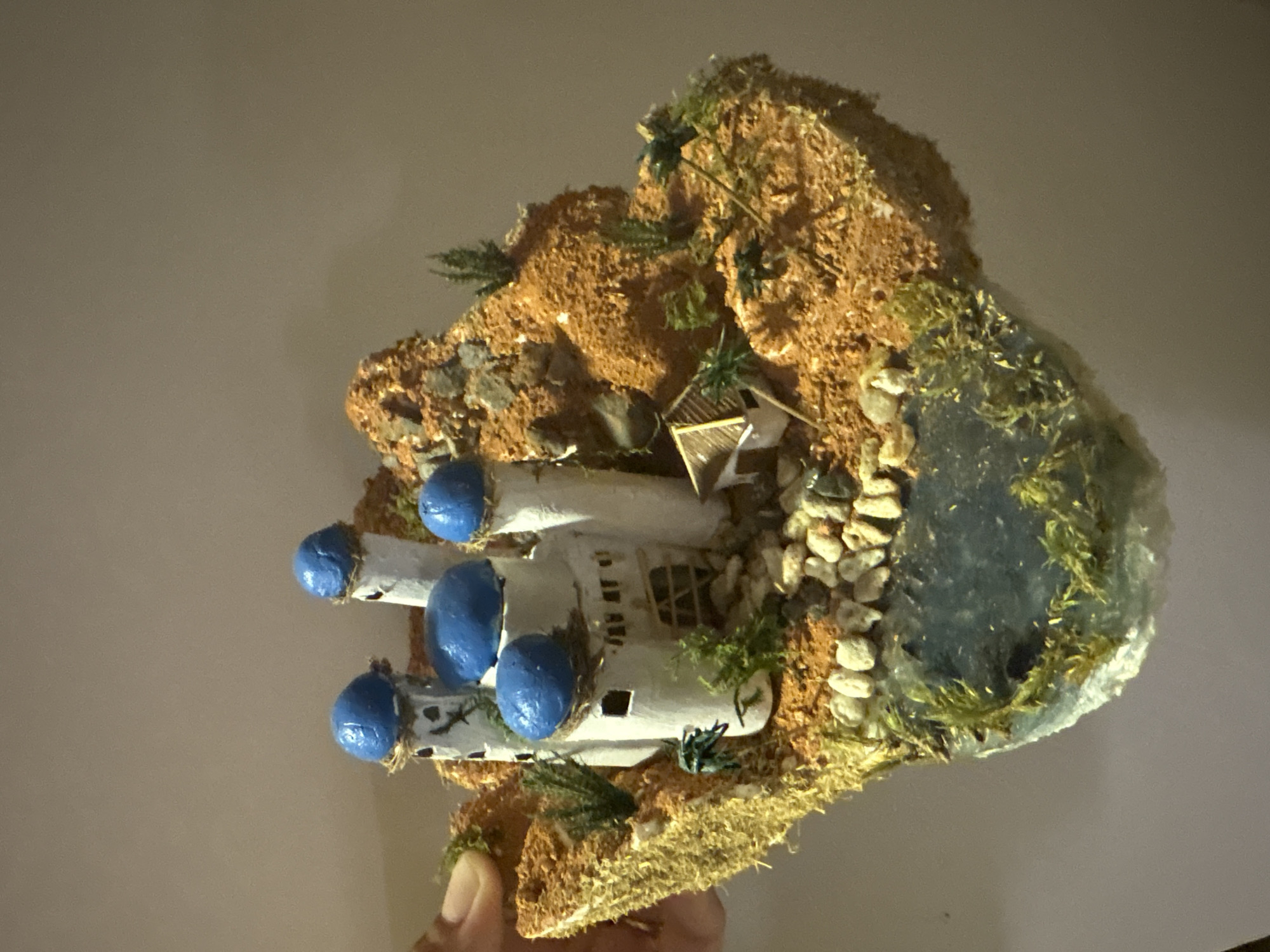 A miniature diorama depicting a fantasy village with white buildings topped with blue domes sits on a textured, orange-brown landscape. The diorama includes a small pond, miniature trees, rocks, and what appear to be tiny figures scattered throughout. The diorama is held by a person's hand on the left side of the image. The background is a plain, light brown wall. The location in the city of Nha Trang Ward, Vietnam, is not visible or inferable from the image.