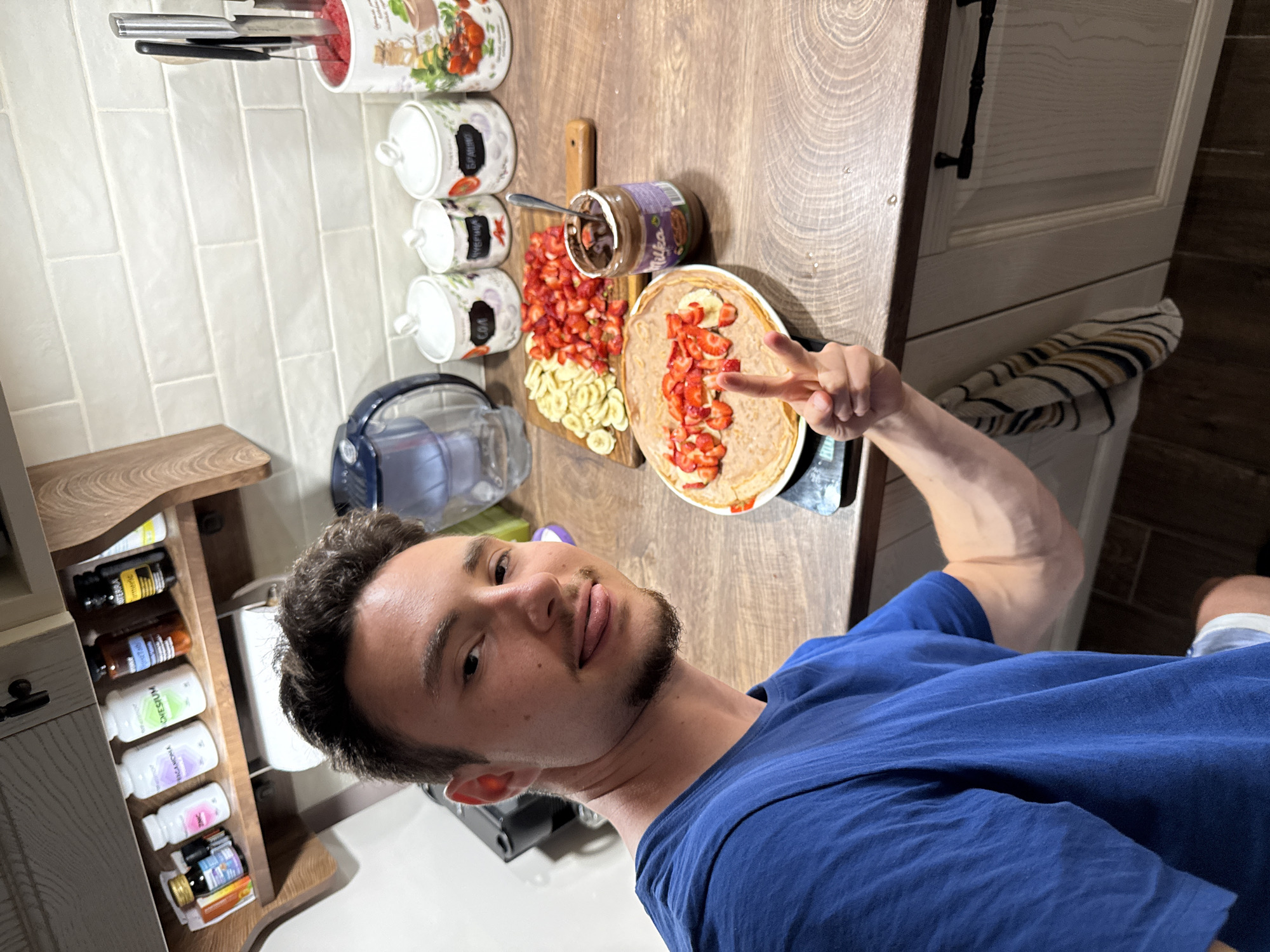 The scene depicts an adult male in a kitchen located in Sofia, Bulgaria, presenting a prepared meal. The man, positioned to the left of the frame, has a light complexion, short dark hair, and a slight beard. He is wearing a royal blue short-sleeved t-shirt and is looking directly at the camera with a subtle smile, his tongue slightly extended. His left hand is raised in a "peace sign" gesture, directed towards a plate of food.
The kitchen features a light-colored tiled backsplash with horizontally laid rectangular tiles and wooden countertops. In the upper left, an open wooden shelf beneath a darker wooden cabinet displays several supplement bottles, identifiable labels include "ZINC," "BERGAMONA," "MAGNESIUM," "OMEGA-3," and "Doterra Turmeric." Below the shelf, a roll of paper towels is mounted.
On the wooden countertop, a food preparation area is set up. A wooden cutting board displays neatly sliced bananas and diced strawberries. A glass jar of "Milka" branded chocolate spread, containing a spoon, is situated next to the cutting board. A white plate, resting on a black digital kitchen scale, holds several crepes or pancakes topped with diced strawberries and sliced bananas.
To the right of the countertop, a blue and clear water filter pitcher is visible. Adjacent to it are three white ceramic storage jars with lids, decorated with botanical patterns, and labeled in Cyrillic as "СОЛ" (Salt), "ЗАХАР" (Sugar), and "БРАШНО" (Flour). Further right, a white knife block with a red interior and a tomato/herb motif holds several kitchen knives. The lower portion of the image reveals white kitchen cabinets with black, wrought-iron style handles, from one of which a striped kitchen towel (blue, yellow, white) is hanging. The floor is covered with dark brown tiles.