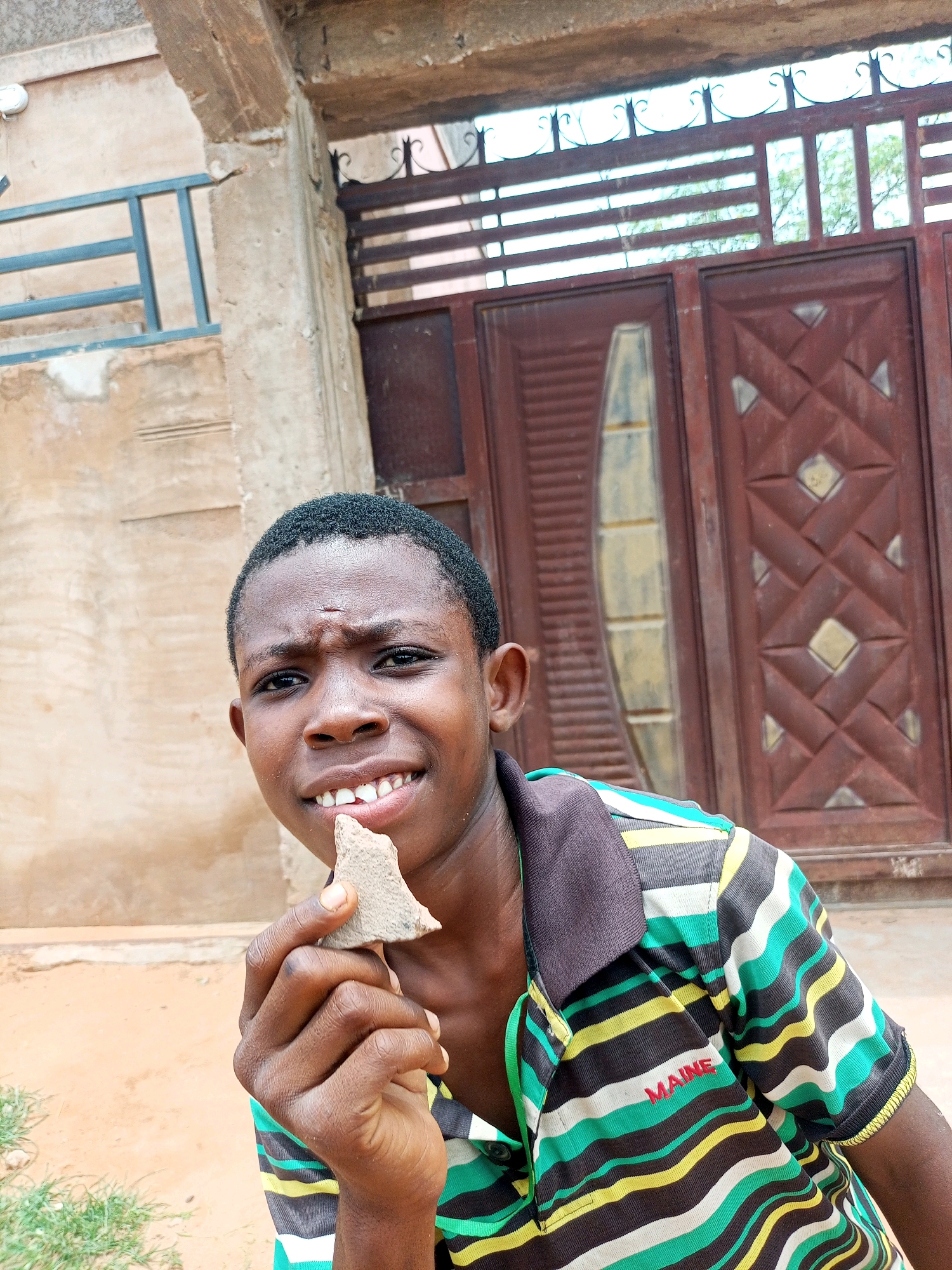 A medium shot shows a young Black male, possibly a boy, looking forward and holding a small, light brown, triangular object to his mouth with his right hand. His left hand is near his mouth, slightly obscured by the object. He has short, dark, curly hair and a dark complexion. He is wearing a collared short-sleeved shirt with horizontal stripes of green, black, yellow, and white. Red embroidered text, partially visible, reads "MAINE" on the left chest area of the shirt. He is smiling, revealing his teeth.
The background consists of a rough, light-colored wall with a textured surface. To the left, a section of a dark metal railing is visible against the wall. Behind him and to the right, a large, dark reddish-brown double door with a geometric design is prominent. The doors have metal accents and a vertical rectangular inset with a light-colored, semi-transparent material. Above the doors, there is a decorative metal grate and beyond that, a bright, hazy sky with visible tree branches. The ground in the foreground is sandy and a patch of green grass is visible on the lower left. The scene appears to be outdoors, likely in a residential or public area. The overall lighting suggests it is daytime.