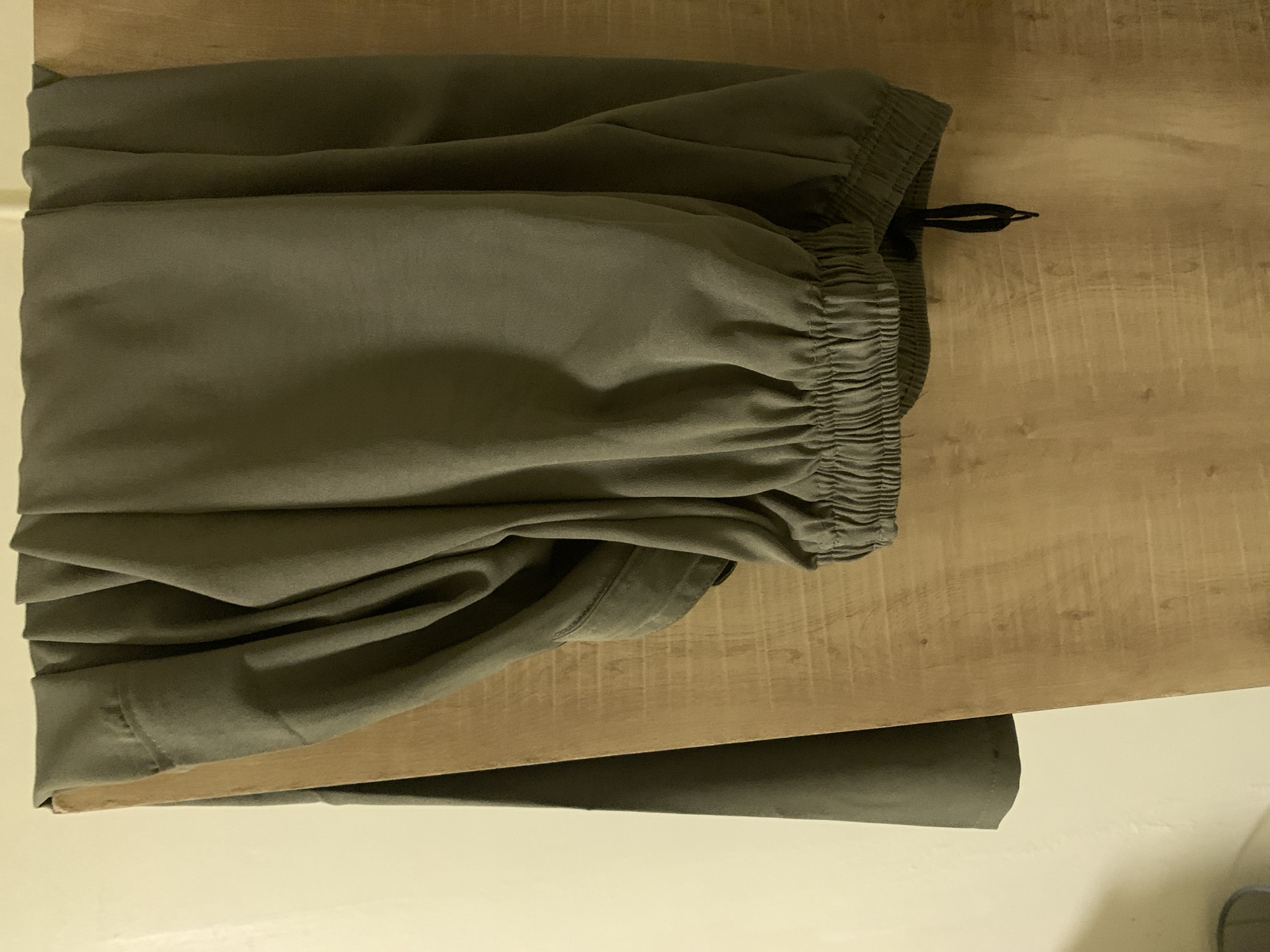 A pair of green pants are hanging on a wooden board. The pants are gathered at the waist and ankles with elastic. A drawstring hangs from the waist. The background is a light-colored wall.