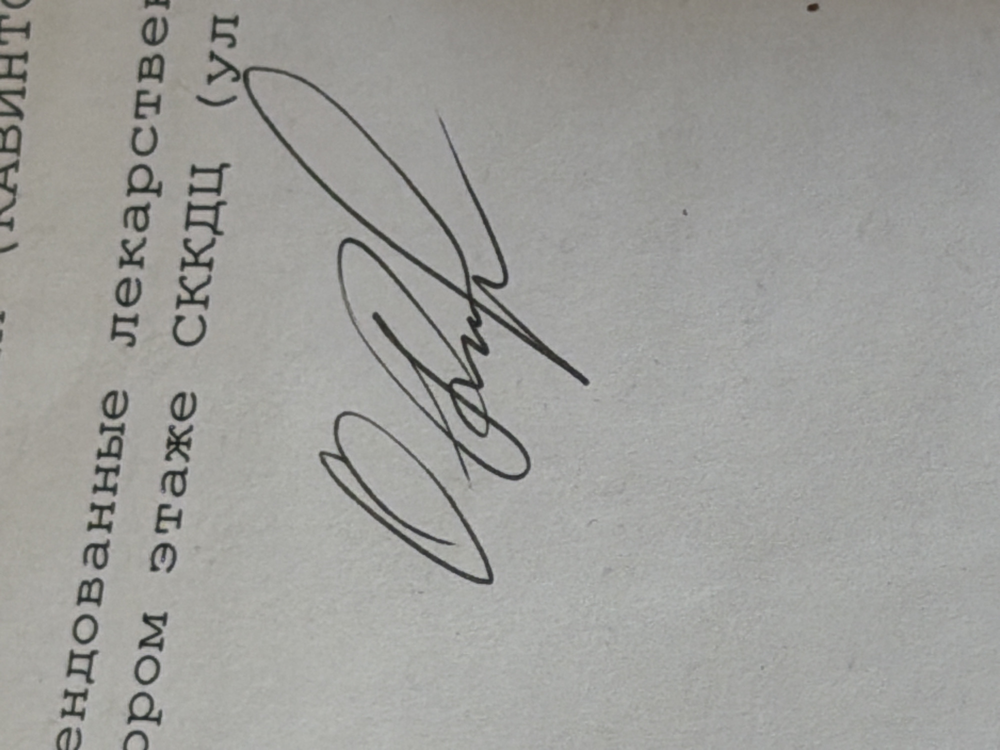 The image presents an angled, close-up view of a document, likely made of paper, featuring both printed Cyrillic text and a prominent handwritten signature in black ink. The paper is light-toned, possibly white or off-white, and shows some subtle texture.
The printed text, arranged vertically in the frame due to the image's orientation, is in Russian. Visible fragments include: "КАВИНТ" (partially visible, potentially part of "КАБИНЕТ" – office/cabinet), "ендованные лекарстве" (suggesting "рекомендованные лекарства" – recommended medicines), "ром этаже" (likely "втором этаже" – on the second floor), and "СККДЦ (ул" (indicating "СККДЦ (улица" – SKKDC (street), with СККДЦ being an acronym, possibly for a medical center).
The signature, bold and expressive, is composed of a series of interconnected loops and flourishes, rendering it largely illegible. It begins with a large, sweeping initial, possibly an 'M' or similar letter, followed by dynamic, continuous strokes. No specific individuals are shown, nor is the time of day evident. The document appears to be an official record or prescription from a medical or administrative context in Moscow, Russia.