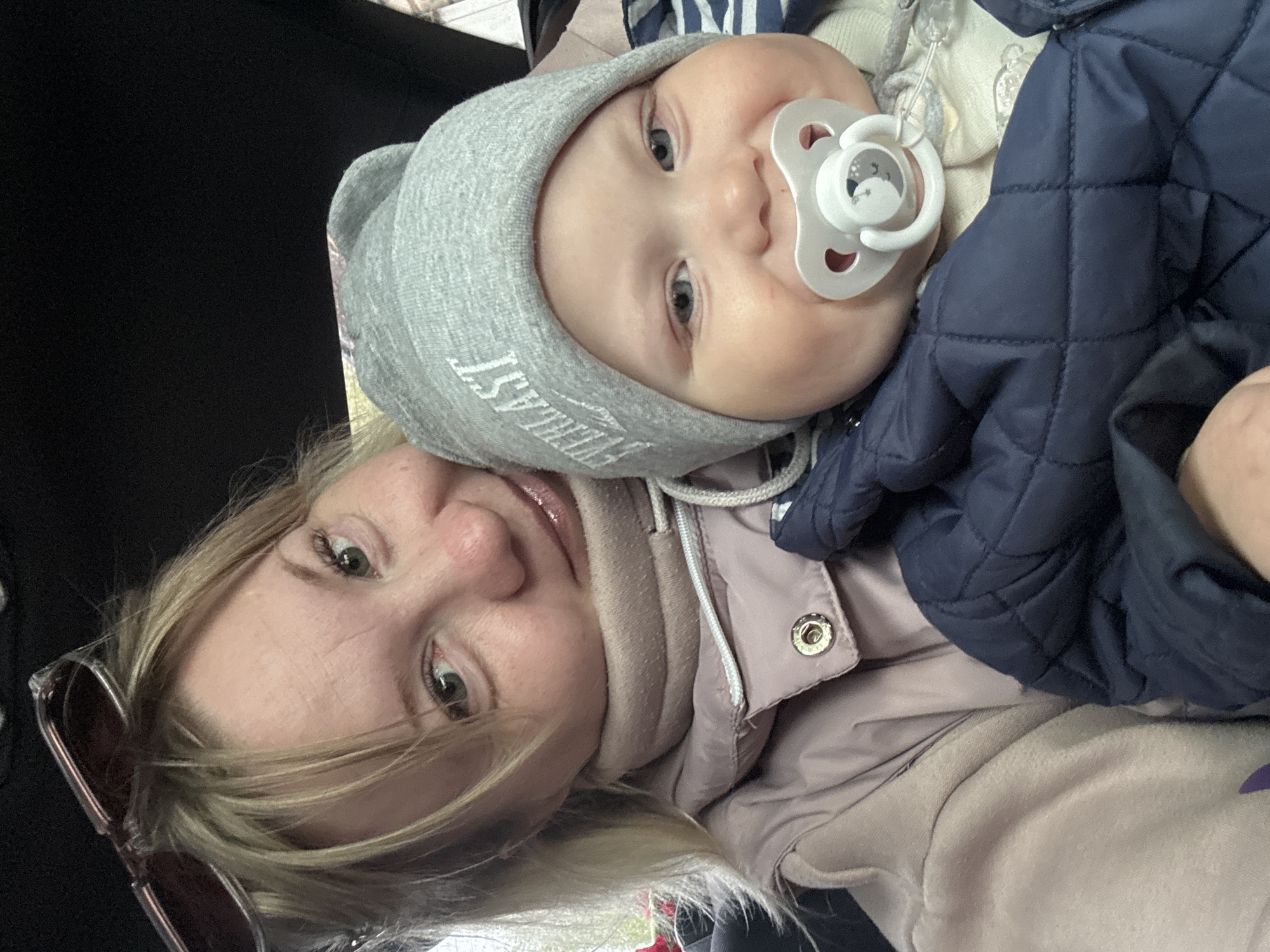 A selfie shows a woman and a baby. The woman has blond hair, fair skin, and green eyes. She is wearing a light pink jacket and sunglasses are visible on her head. The baby is wearing a gray hat with the word "EVERLAST" on it and a navy blue quilted jacket. The baby has a pacifier in their mouth. The background is dark. The location is likely the city of Chaikovsky, Russia based on user prompt.