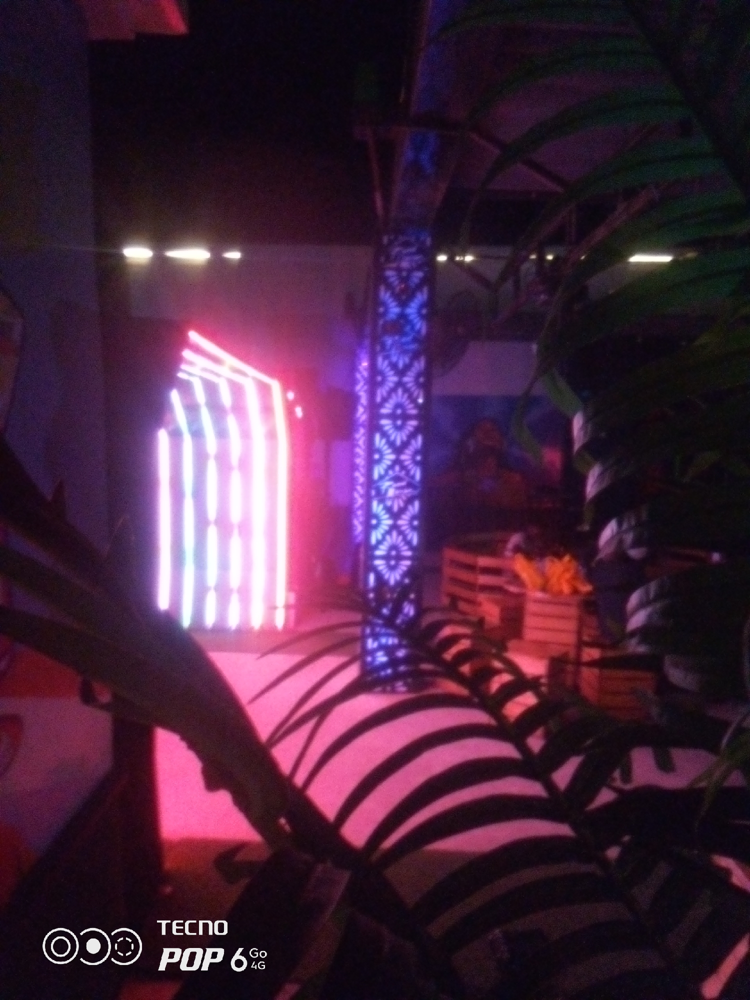 The image depicts a dimly lit indoor scene, possibly an entertainment venue, with vibrant neon lighting. In the foreground, large dark green palm fronds cast shadows on a pink and black striped surface. To the left, a structure with vertical pink and white neon lights creates an archway effect. In the background, a tall, ornate pillar glows with blue and purple patterns. A wooden crate filled with orange objects is visible behind the pillar, and several stacked tires are present to the right. The "TECNO POP 6 GO 4G" logo is present in the bottom left corner of the image. There are no discernible people or specific identifiable landmarks of Howayi, Nigeria in the image.