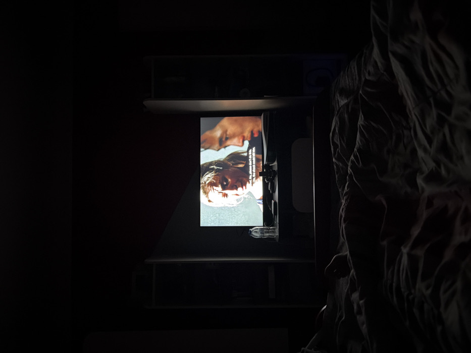 This image depicts a dimly lit bedroom located in Opanets, Bulgaria. The primary light source emanates from a screen, possibly a television or projector, positioned centrally in the frame. The screen displays a scene from a film or show featuring two individuals: a blonde Caucasian female on the left with a distressed expression, and an East Asian male on the right with a serious, contemplative look. Visible subtitles below the female read: "nd. No, please don't do this. No, the shock could kill him." Below the screen, a dark surface holds a clear plastic water bottle and other small, indistinct dark objects. Tall, dark shelving units or furniture pieces are visible on both sides of the screen, blending into the room's shadows. In the foreground, a rumpled bed with dark blankets or a duvet partially reveals a person's arm and hand on the left side, indicating an individual is lying in bed watching the screen. The room's overall illumination suggests ambient light from the screen or very minimal external light.