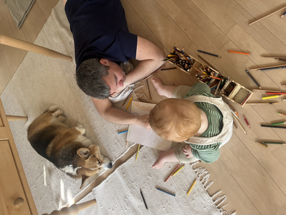 An indoor scene in Hohenbrunn, Germany, depicts a male adult and an infant engaged in a coloring activity on a light-colored wooden floor. A Pembroke Welsh Corgi dog is lying nearby on a textured, light-colored rug.
The adult male, exhibiting short, graying hair, is positioned on his left side on the rug, supporting his head with his left hand which rests on a striped cushion. He is wearing a dark blue t-shirt. His right arm is extended towards two sheets of white paper placed on the floor. One sheet displays a light outline sketch.
The infant, observed from behind, has reddish-brown hair and is seated on the wooden floor, facing the adult male and the paper. The infant wears a green and white striped long-sleeved shirt beneath light-colored overalls.
A wooden pencil holder, containing multiple compartments filled with colored pencils, is situated on the floor to the right of the infant. Numerous individual colored pencils and crayons are scattered across the wooden floor and partially on the rug, surrounding the participants.
The tri-color Corgi is lying prone on the rug to the left of the adult male, with its head resting downwards.
Partial views of a light-colored wooden table's legs and underside are visible in the upper left quadrant of the frame, resting on the rug. Natural light casts subtle shadows on the rug from an indeterminate source.