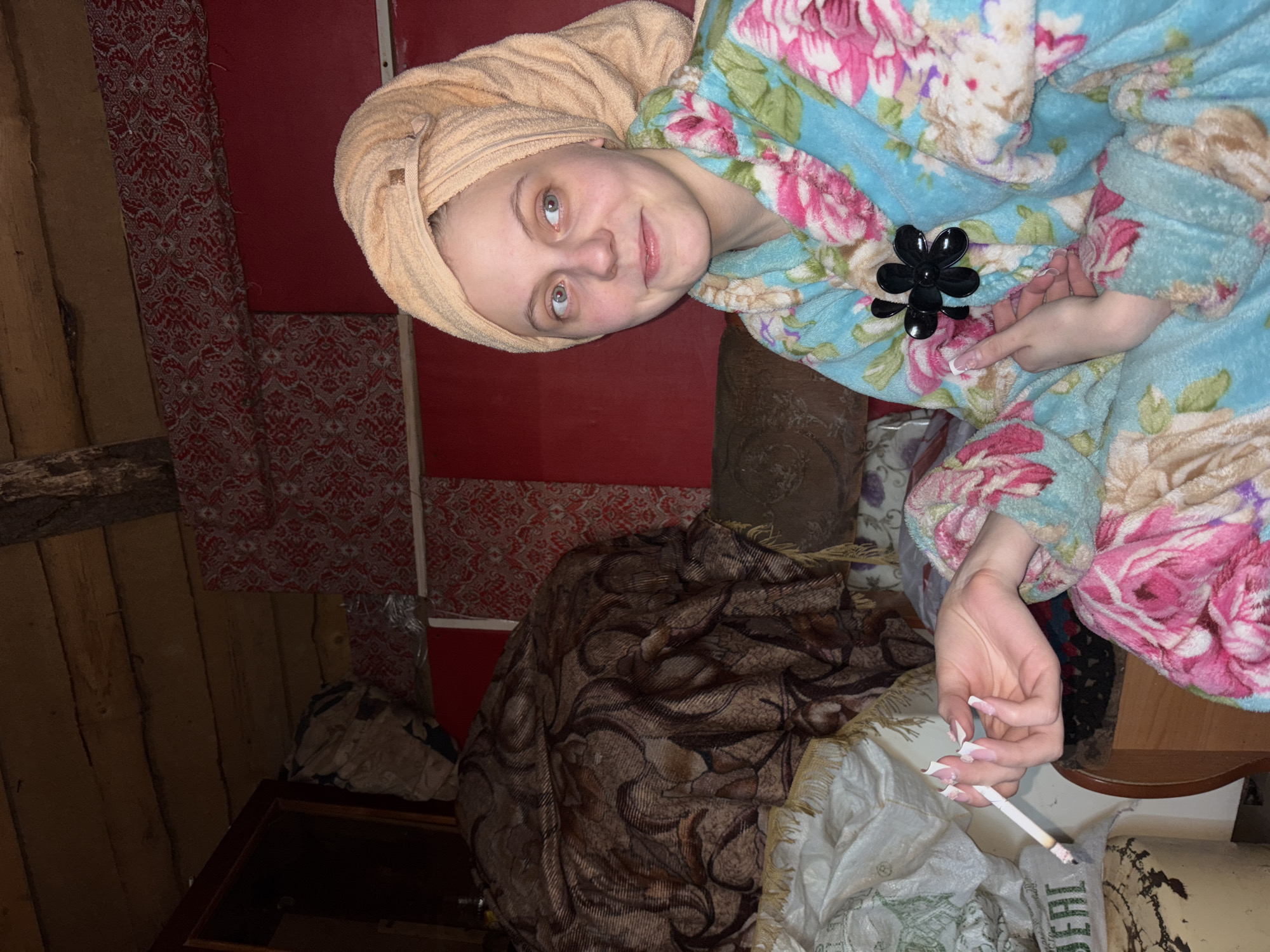 The image features a young woman, likely indoors, wearing a peach-colored towel wrapped around her head and a blue floral bathrobe. She is holding a lit cigarette in her right hand. The background shows a room with rustic elements, including wooden walls, a red wall covering with a patterned design, and furniture draped with a brown patterned blanket and some white plastic bags. The text "ВЕЩЬ" is visible on a bag near the cigarette. The location cannot be determined with certainty to be in the city of Фоки, Russia, without additional information or context.