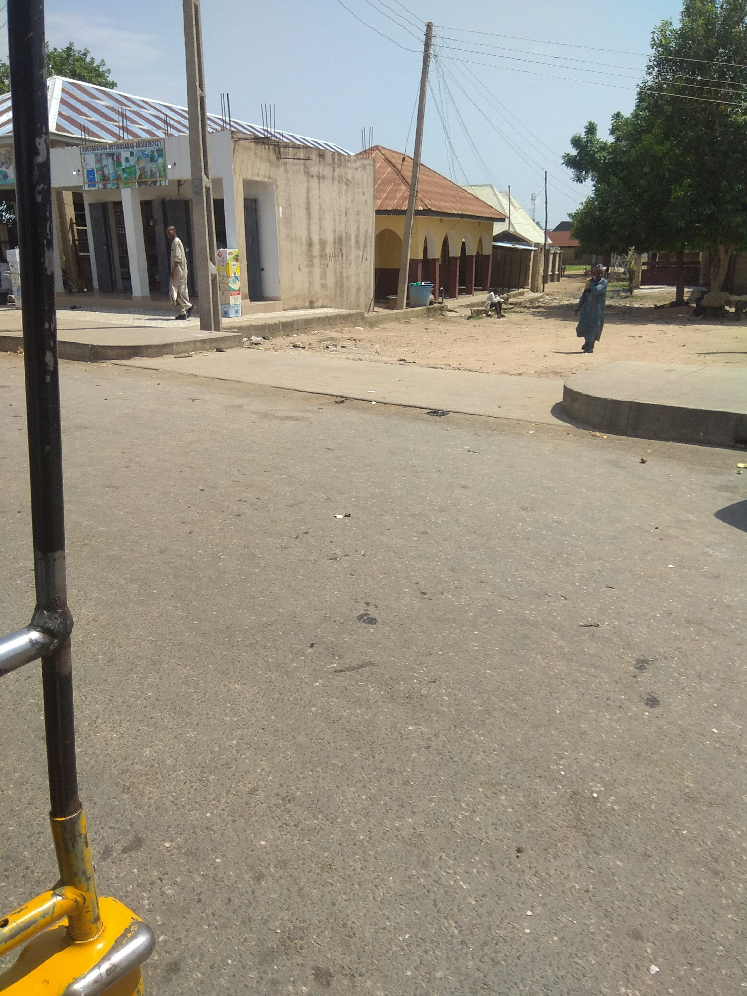 The image captures a street scene in Jalingo, Nigeria, with a focus on a building complex and the surrounding environment. In the foreground, a portion of a yellow and silver railing is visible, suggesting the perspective is from a vehicle or elevated structure. The street is paved with asphalt, showing signs of wear and scattered debris.
To the left, a building with a corrugated metal roof, painted with red and white stripes, stands partially constructed. It features white columns supporting an overhang and several doorways. Signs are affixed to the building's facade, one displaying text that includes "0803567644-08118054845-08149782421". A man wearing a light-colored garment walks in front of this building. Next to him, several stacked boxes are visible.
Adjacent to this structure is another building with a beige, rough-textured wall and a doorway. Further down the street, more buildings with reddish-brown, corrugated metal roofs are visible, some with arched openings and dark brown trim. A woman in a long blue garment is seen walking on the dirt and paved area to the right, away from the viewer. A large, leafy green tree stands to the right of the street, casting shadows. Power lines are strung across the sky, supported by utility poles. The overall environment appears to be a commercial or residential area with ongoing construction and a modest level of activity. The sky is clear and blue.