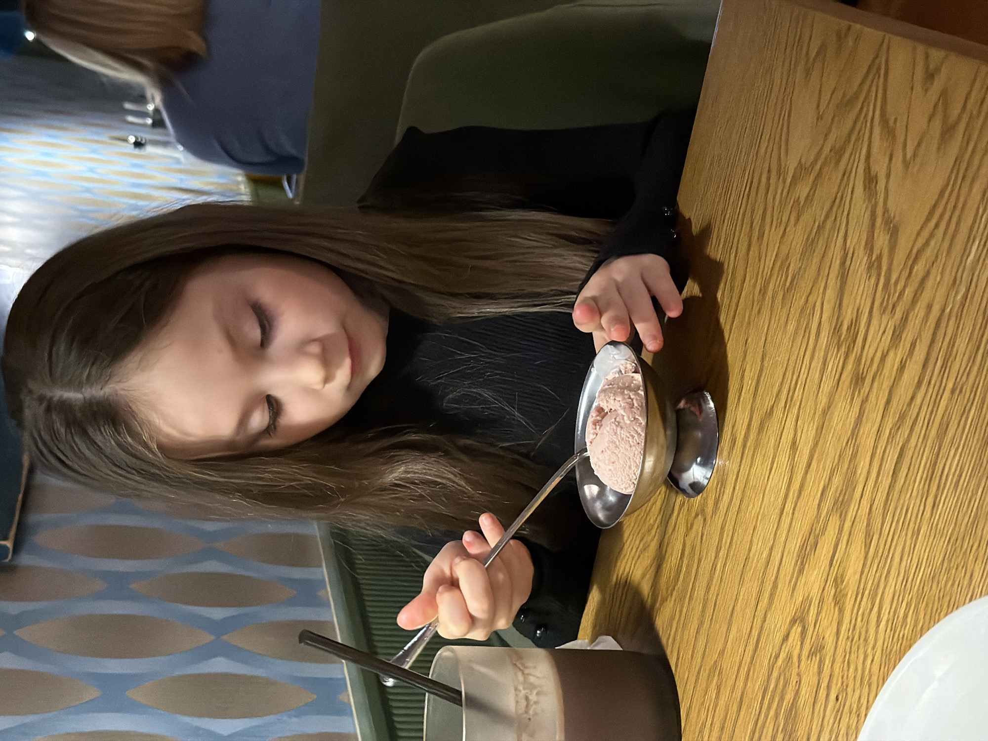 A young girl with long brown hair is seated at a wooden table in what appears to be a restaurant. She is wearing a black long-sleeved shirt and has her eyes closed, appearing contemplative. In front of her, there is a metal bowl containing what seems to be strawberry ice cream. She holds a spoon in her right hand, partially inside the bowl. To the lower left of the bowl, there is a tall glass containing a brown beverage, possibly a milkshake or hot chocolate, with a straw inside. The background includes a wall with a blue and beige patterned design and another person with blonde hair wearing a blue shirt sitting at a nearby table. The photo was taken in Perm, Russia.