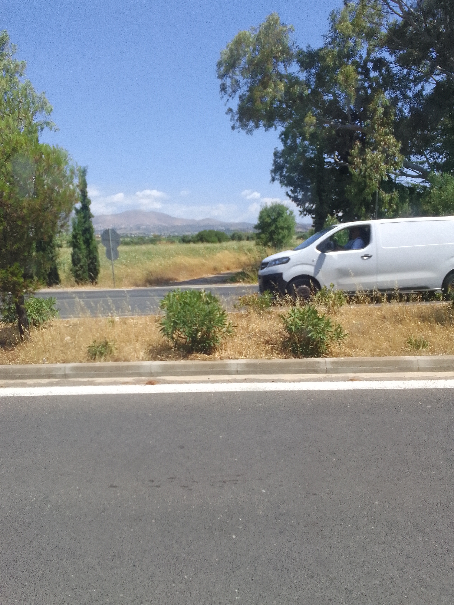 A white commercial van is driving on a road with dry grass and vegetation on the side. In the background, there are hills and trees under a clear blue sky. The scene appears to be in a rural or suburban area, possibly near Spata-Loutsa Municipal Unit, Greece.