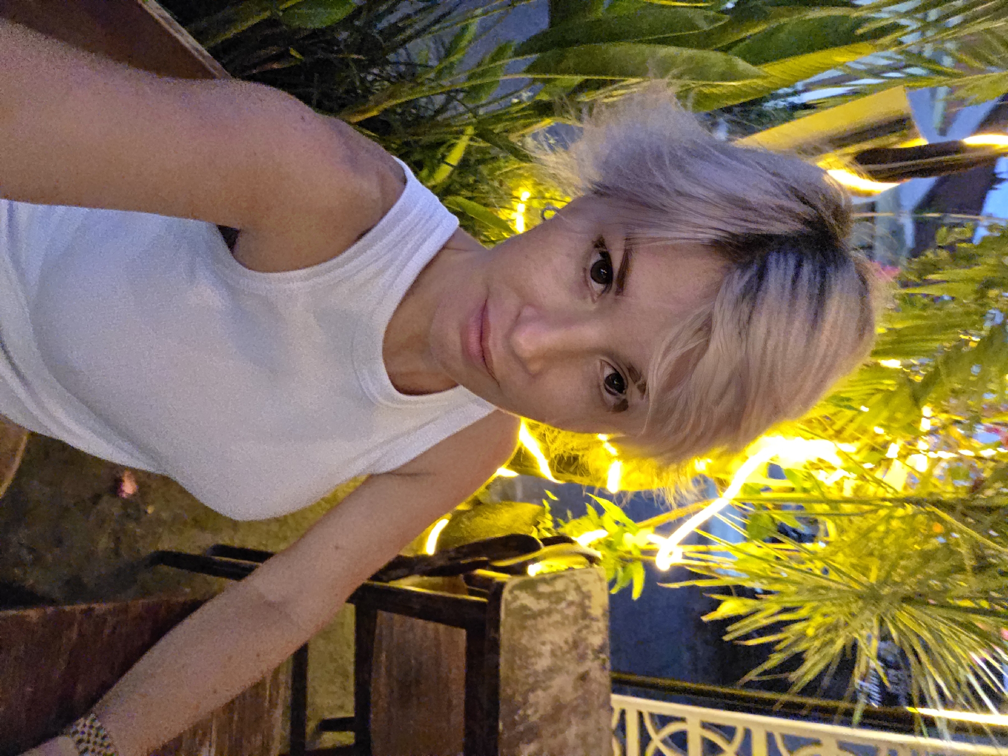 A woman with short, light blonde hair and a white tank top takes a selfie outdoors at night. She is sitting at a wooden table and chair. Behind her, lush green plants and bright yellow string lights create a festive atmosphere. The background suggests a tropical setting, possibly a restaurant or bar, consistent with the location of North Kuta, Indonesia.