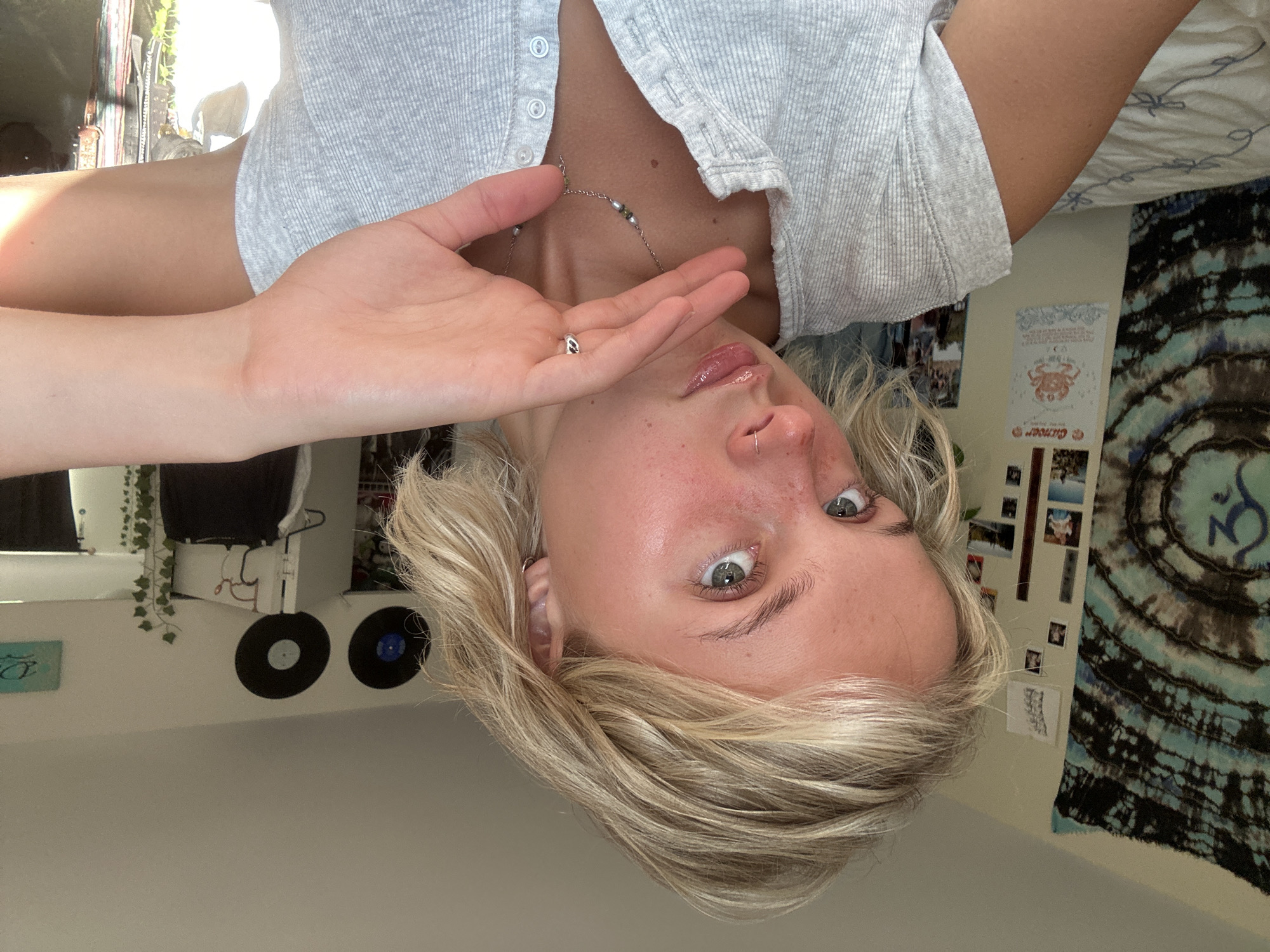 A young woman with short, blonde hair is shown from the chest up, looking directly at the camera. She is wearing a grey, ribbed shirt. Her right hand is raised to her face, partially obscuring it. She has a nose ring and a small ring on her right ring finger. A necklace with small beads is visible on her chest. The background appears to be a bedroom, with a tapestry on the left wall and framed pictures and vinyl records on the wall behind her. There is a bed with white bedding visible at the bottom left of the image. The lighting suggests it is daytime, with sunlight coming from the right side of the frame. The image was taken in Whistler, Canada.