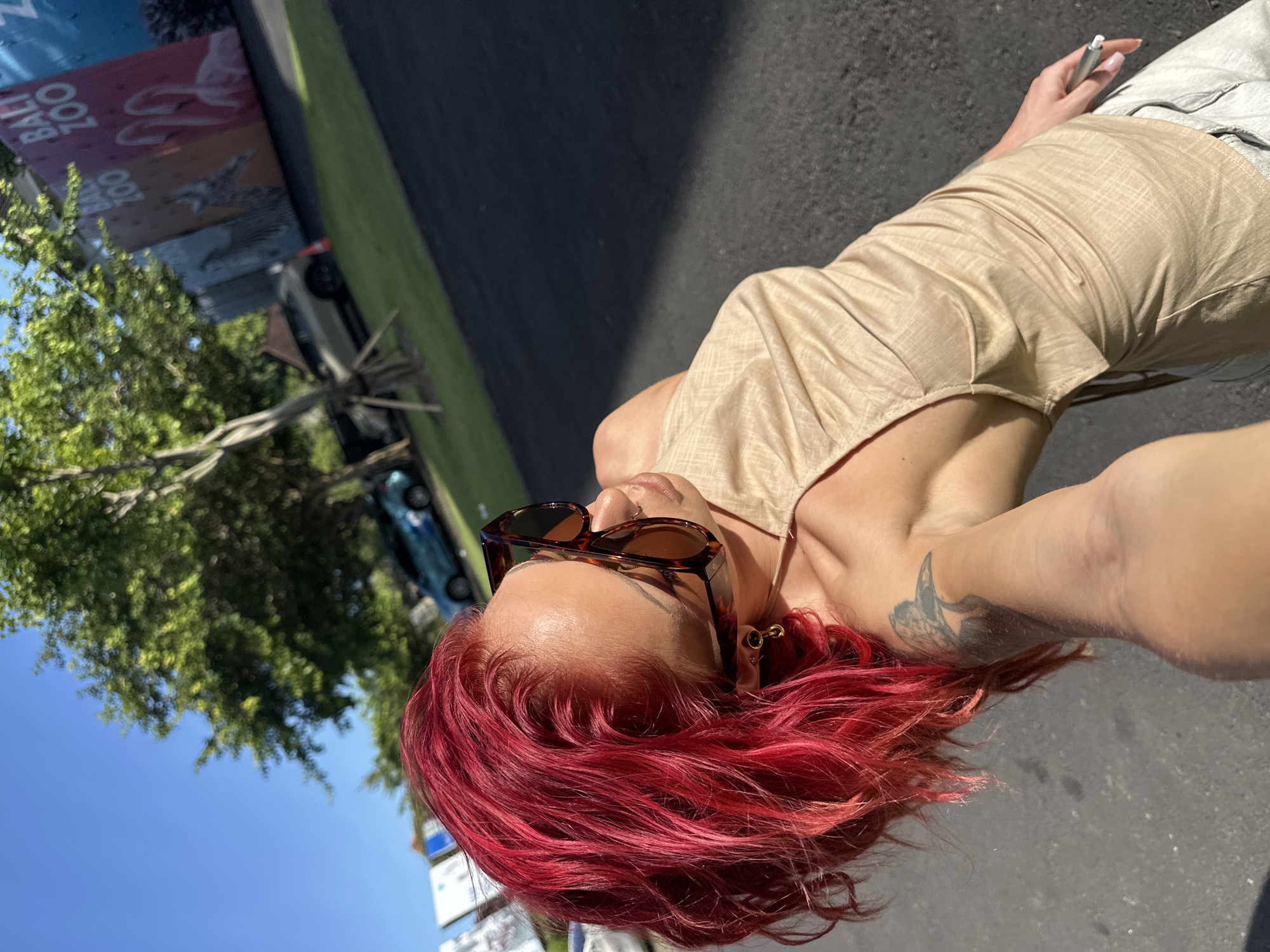 A selfie shows a person with red hair, wearing brown sunglasses and a beige shirt, standing on an asphalt surface near a sign for the Bali Zoo in Sukawati, Indonesia. A car and trees are visible in the background under a blue sky. The person holds a pen in their hand. A tattoo is seen on their arm.