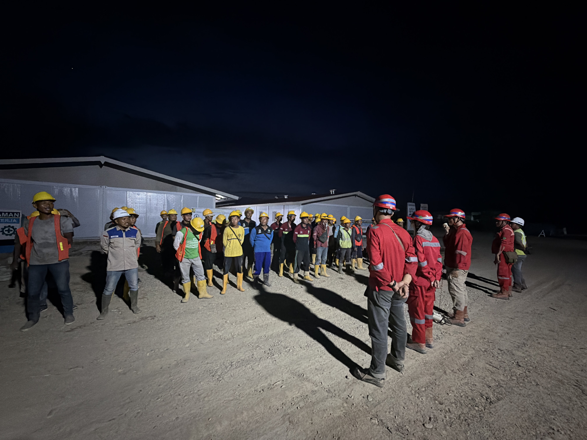 The image depicts a group of workers gathered outdoors in Simpangempat, Indonesia, at night. Most of the workers are wearing yellow hard hats, while others wear red or white hard hats. Some wear reflective vests. A few are in red jumpsuits. Most are wearing boots. The group is standing on a dirt surface with some visible ruts. There are low buildings with white walls visible in the background. The sky is dark, indicating nighttime. Shadows are cast by the workers.