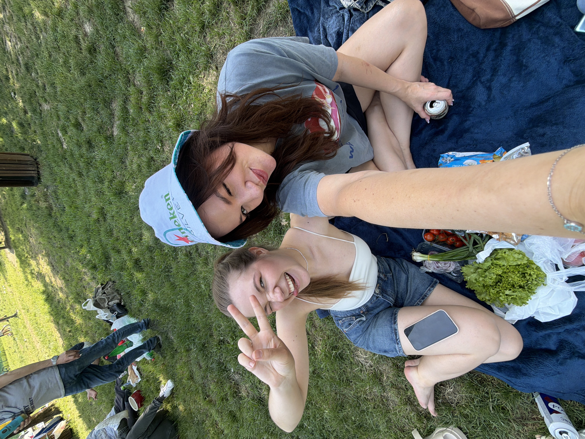 Two young women are taking a selfie outdoors, likely at a picnic. They are sitting on a blue blanket on a grassy area. The woman closer to the camera is making a peace sign and has a phone on her leg. There is food visible near them, including tomatoes, onions, and lettuce. There is a can of beer near her feet. Behind them and to the left, there are other people and belongings scattered on the grass. Given the items included, this photo was taken in Cukarica Urban Municipality, Serbia.