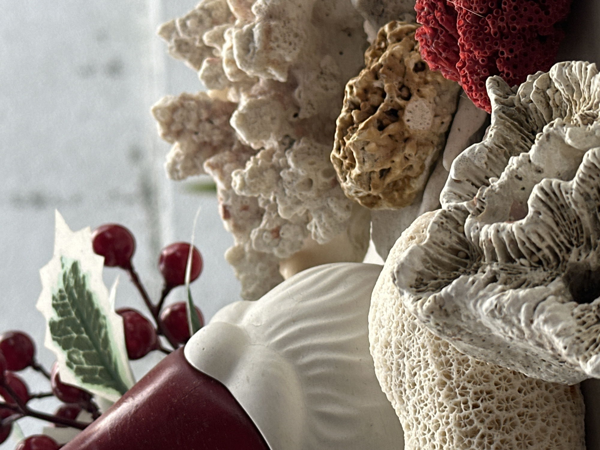 The image shows a close-up of a decorative arrangement. It includes pieces of white and beige coral, a piece of red sponge, artificial holly berries and leaves. Part of a red and white figurine is visible. The composition is tightly cropped, and the setting is undifferentiated. There is no indication of the location being North Kuta, Indonesia.