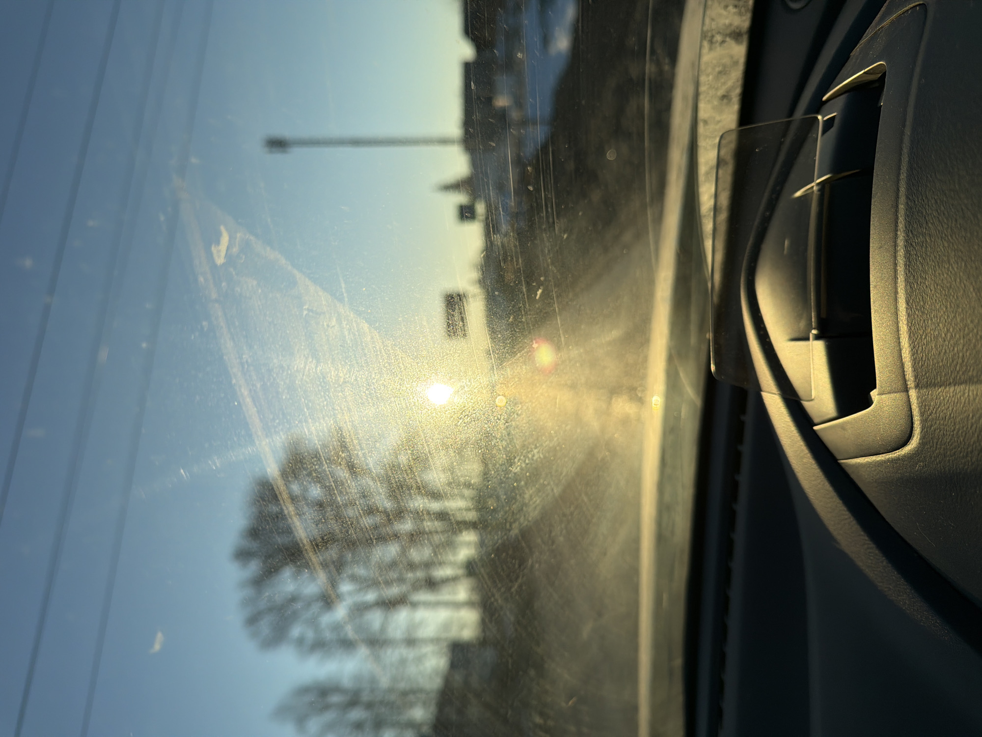 The image depicts an exterior scene viewed through the dirty and scratched windshield of a vehicle in the city of Chaikovsky, Russia. The low-angle sun, positioned slightly right of center, causes significant glare and lens flare across the windshield, dominating the central and right portions of the frame. The sky is clear and blue. To the left, silhouettes of bare deciduous trees are visible against the sky, with multiple power lines running horizontally across the upper left. To the right, indistinct distant structures, including a tall, slender mast or tower, are vaguely discernible through the glare and atmospheric haze; some patches of snow are also apparent on the ground. The foreground shows the dark, textured dashboard of the vehicle, with a heads-up display unit, comprising a dark base and a clear, rectangular screen, positioned centrally. No individuals are visible.