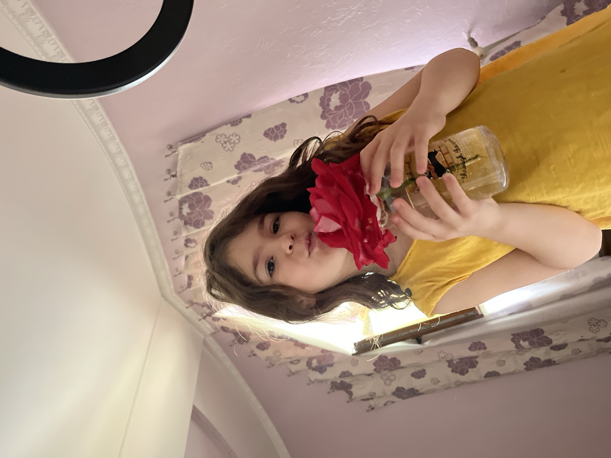 An indoor, vertically oriented photograph, taken in Tarnivtsi, Ukraine, captures a young girl from an overhead perspective. The girl, with long, wavy dark hair and light skin, looks directly towards the camera with a soft expression, appearing content. She is wearing a mustard yellow top or dress.
Her hands cradle a clear glass bottle or jar containing a large, vibrant red rose, which she holds near her face, possibly smelling it. The rose appears to be artificial or made of fabric. The glass bottle features a dark silhouette design, possibly a person or a ship, along with some cursive text above it, which is difficult to decipher but may include "Black Horse".
The background reveals a domestic setting, likely a bedroom or living space. A light-colored curtain with a repeating purple floral pattern is visible behind the girl. The walls are predominantly white, and a light pink ceiling with decorative trim is also visible. In the upper left corner of the original, rotated image, a black circular object, possibly a ring light or part of a lamp, is partially seen. A thin, dark rod or frame, potentially part of furniture or a window frame, runs horizontally in the background. The bright lighting suggests it is daytime. The image appears to be taken from a high angle, looking down at the girl.