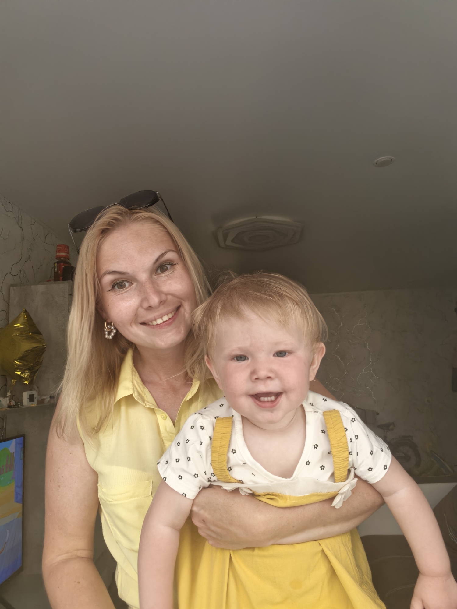 A medium shot captures a fair-skinned woman with blonde hair and a bright yellow sleeveless shirt holding a baby. The woman is smiling at the camera, and sunglasses are perched on her head. The baby, also fair-skinned with blonde hair, is looking at the camera and smiling, with its mouth open to reveal teeth. The baby is wearing a white t-shirt with black floral patterns and yellow suspenders, and a yellow dress. The background shows a wall with a textured pattern and a golden balloon. The scene appears to be indoors.