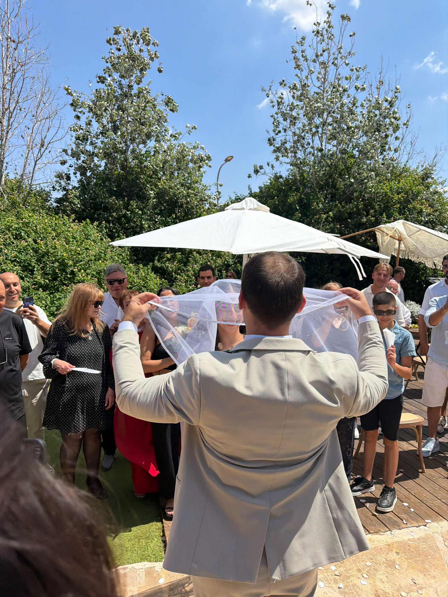 In Kadima, Israel, a wedding ceremony takes place outdoors. The foreground is dominated by a man in a light-colored suit, seen from behind, holding a white veil aloft. Behind him, a bride, her face partially obscured by the veil, is visible. A crowd of guests, dressed in a variety of attire, observes the ceremony. They stand on a wooden deck and a patch of green artificial turf. The background features lush green bushes and trees, with a clear blue sky overhead. Two large white umbrellas are present, suggesting an outdoor event. The atmosphere appears celebratory.