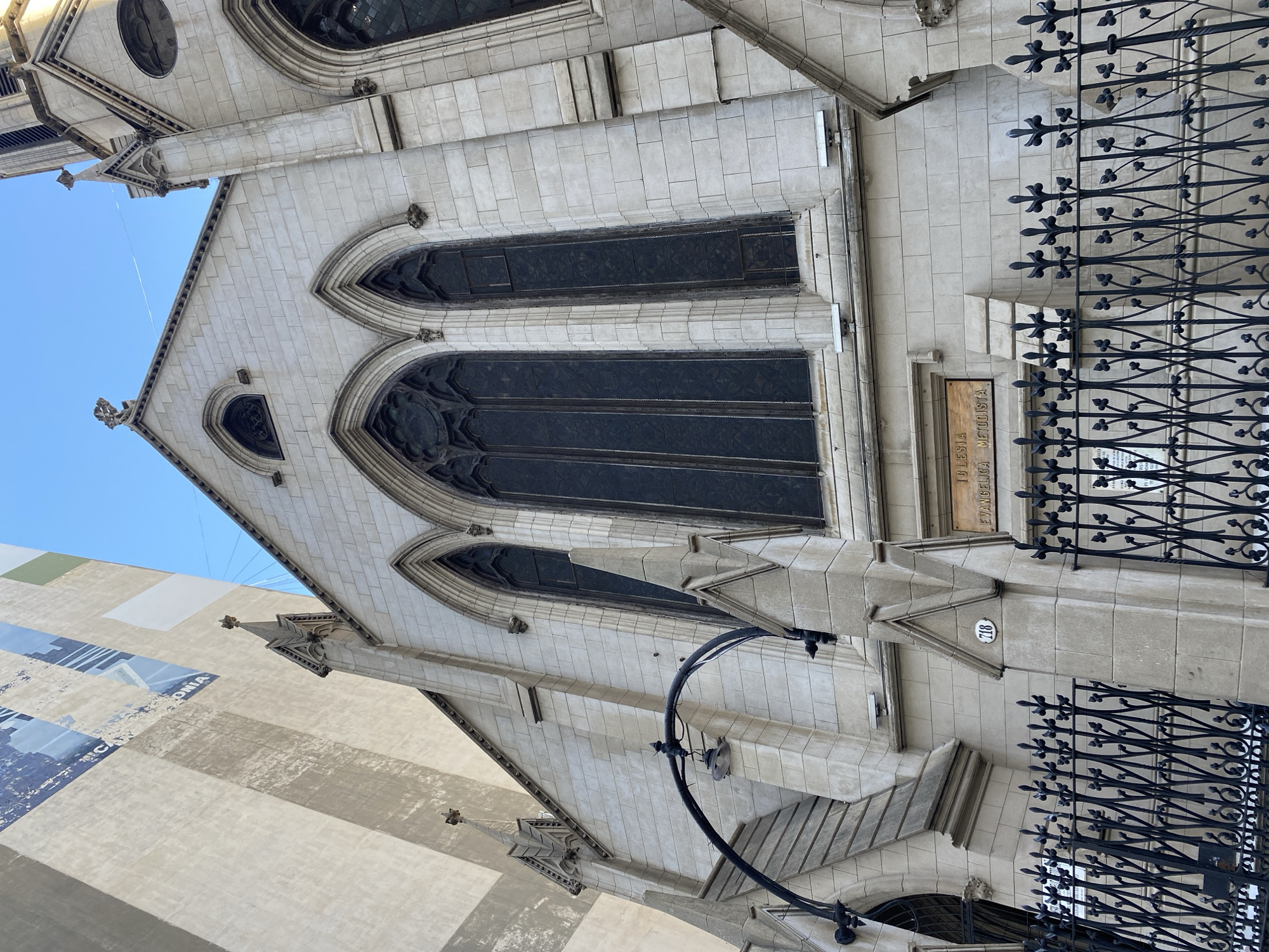 The image depicts the facade of the Iglesia Metodista Evangélica Central, located in Buenos Aires, Argentina. The building is constructed of light-colored stone with Gothic architectural elements including arched windows, spires, and decorative stonework. Black iron fencing runs along the front of the building. A sign on the facade identifies the church. Street lamp is seen near the bottom of the image. A neighboring building's painted mural is visible in the background.