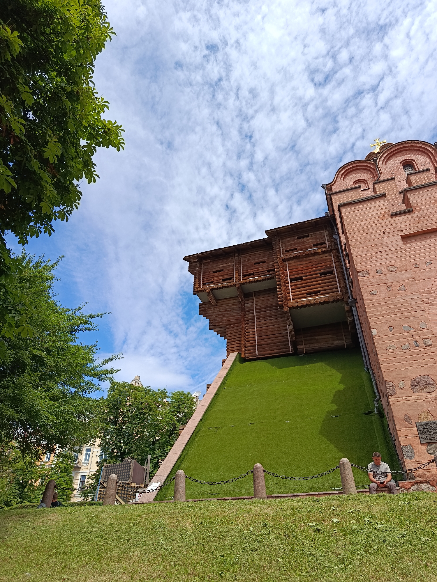 A man sits on a stone bench, looking down at his phone, in front of a historic brick structure in Kyiv, Ukraine. The structure, which appears to be part of the Golden Gate of Kyiv, is made of reddish-brown bricks and features a wooden facade on an angled green surface. Lush green trees frame the left side of the image, and a cloudy blue sky fills the upper portion. A chain fence with stone posts separates the grassy area from the structure.