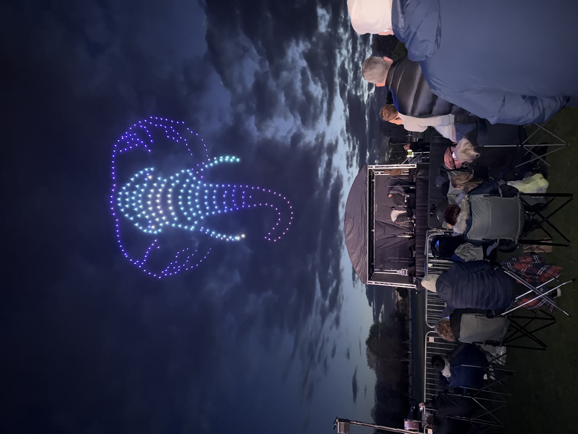 The image shows a nighttime event in South Norfolk, United Kingdom. A large drone formation display in the sky depicts a jellyfish illuminated with blue, purple, and white lights. Below, people are seated in folding chairs, viewing the spectacle. A stage is visible with equipment. The sky contains clouds, and ground level vegetation is present.