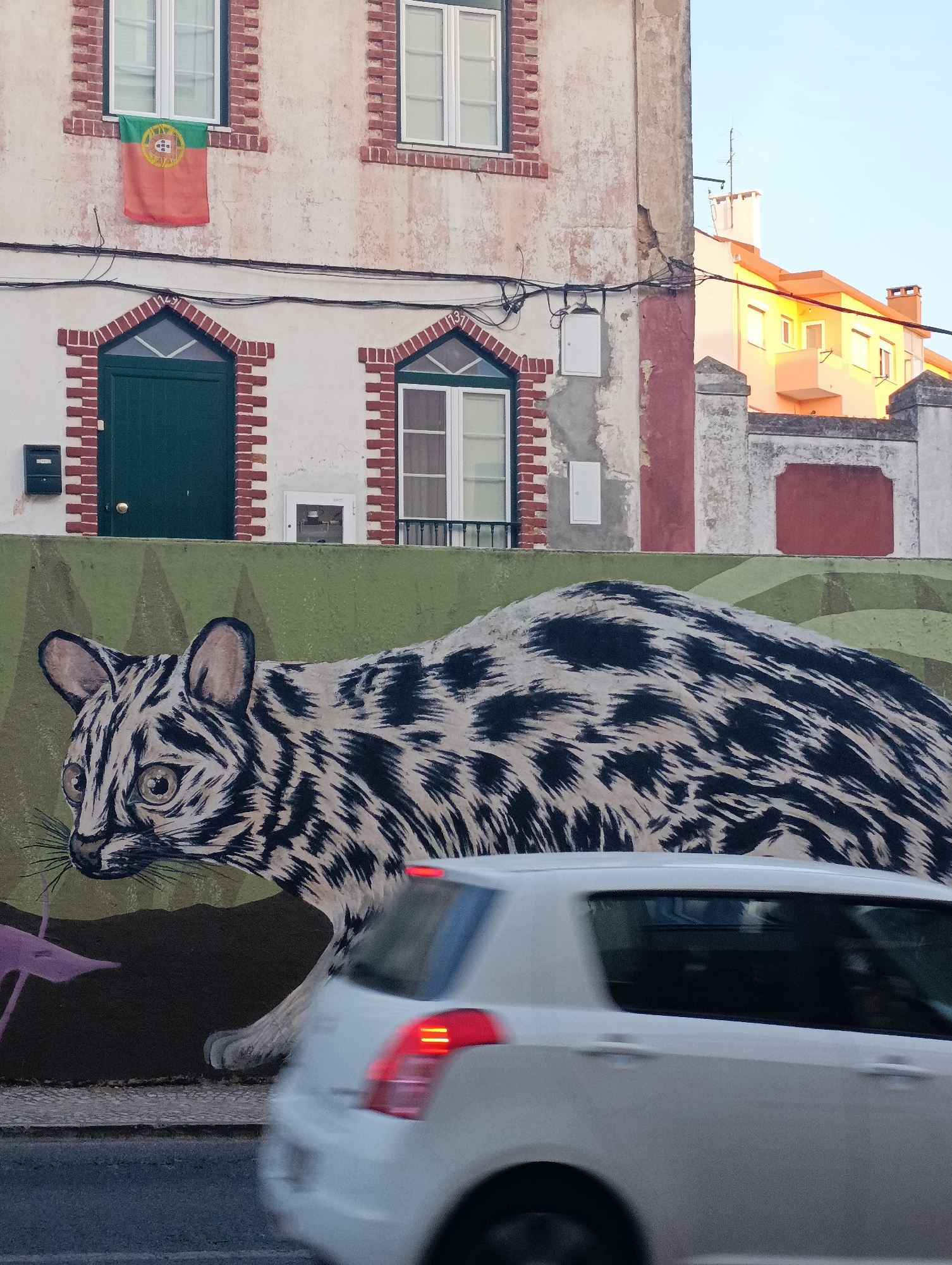 The image depicts a vibrant mural of a leopard-like creature adorning a green wall in Setúbal, Portugal. The mural, painted in black and white with striking detail, covers a significant portion of the wall. Above the mural, the facade of a building is visible, featuring red brick accents around two green doors and windows. A Portuguese flag hangs from one of the windows. In the foreground, a white car is captured in motion, its tail lights blurred, suggesting it is driving past the mural. The background shows a bright yellow building under a clear blue sky. The scene is a lively streetscape, characteristic of a European city with an artistic flair.