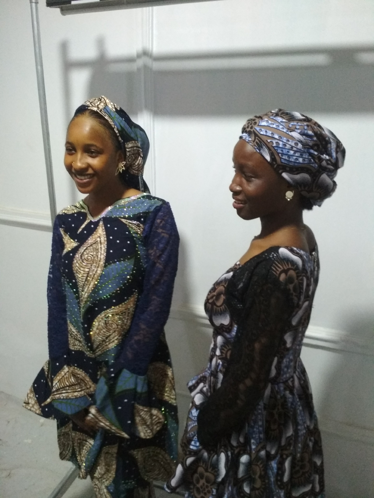 Two women are standing indoors. The woman on the left is smiling and wearing a patterned dress with lace sleeves and a head covering. She is also wearing earrings. The woman on the right is wearing a similar patterned dress and head covering with earrings, and she is also smiling. They are standing in front of a white wall with a lighting fixture at the top. The location is Unknown, Unknown.