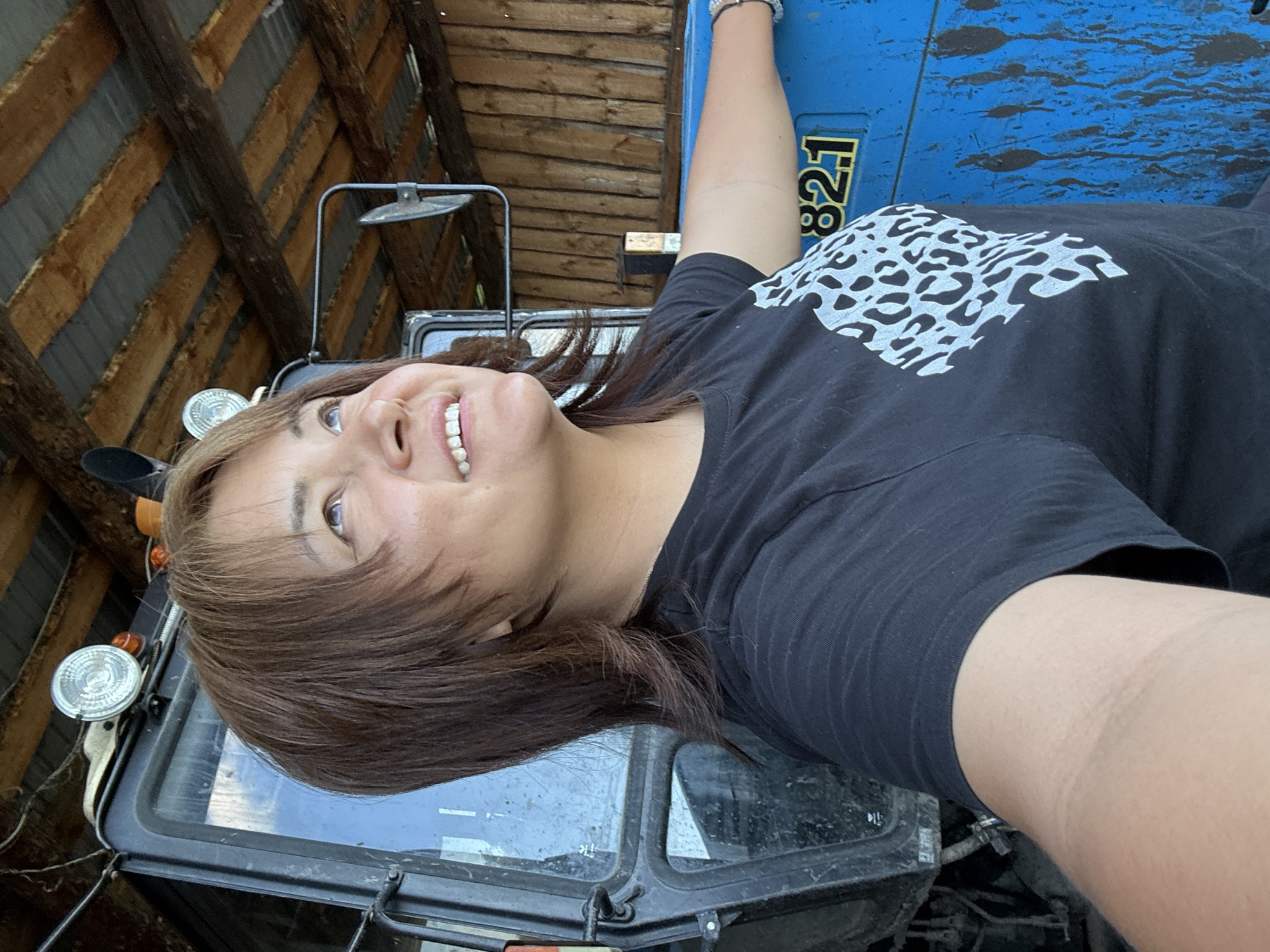 A woman with shoulder-length, layered brown hair, parted to the side, is pictured from the chest up, looking upwards to her right. She is wearing a black t-shirt with a white leopard print design on the left side of her chest. Her right arm is extended to her right, resting on a blue tractor with yellow numbers "821" visible. The tractor is situated inside a wooden structure with a corrugated metal roof supported by wooden beams. The background consists of wooden walls. The scene appears to be outdoors or in a semi-enclosed structure, possibly a farm or rural setting.