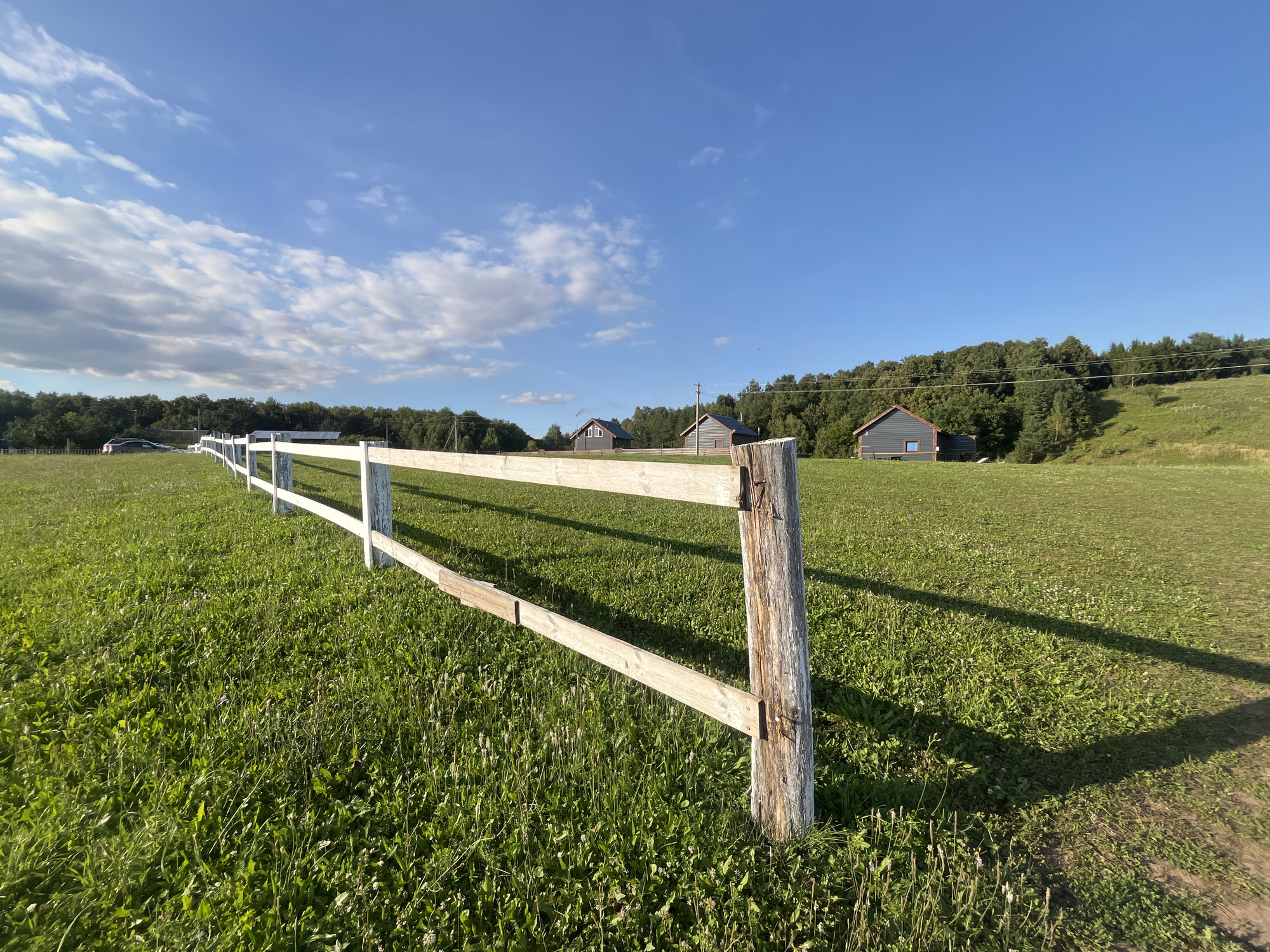 This image depicts a rural scene in Srednie Pinyachi, Russia, characterized by a lush green field under a bright blue sky with scattered white clouds. In the foreground, a weathered wooden fence post supports a white railing that extends diagonally across the frame, casting a long shadow onto the grass. The field itself is covered in dense green grass, interspersed with small weeds and wildflowers, and appears to be sloped gently upwards in the distance.

Further back, several small, dark-colored wooden houses with reddish-brown roofs are visible. These houses are nestled amongst a dense line of trees that form the backdrop of the scene. To the left, a dark-colored car is parked on what appears to be a dirt path or road. The overall impression is one of a tranquil, natural landscape with rustic human habitation. The lighting suggests it is daytime, likely late afternoon due to the long shadows.