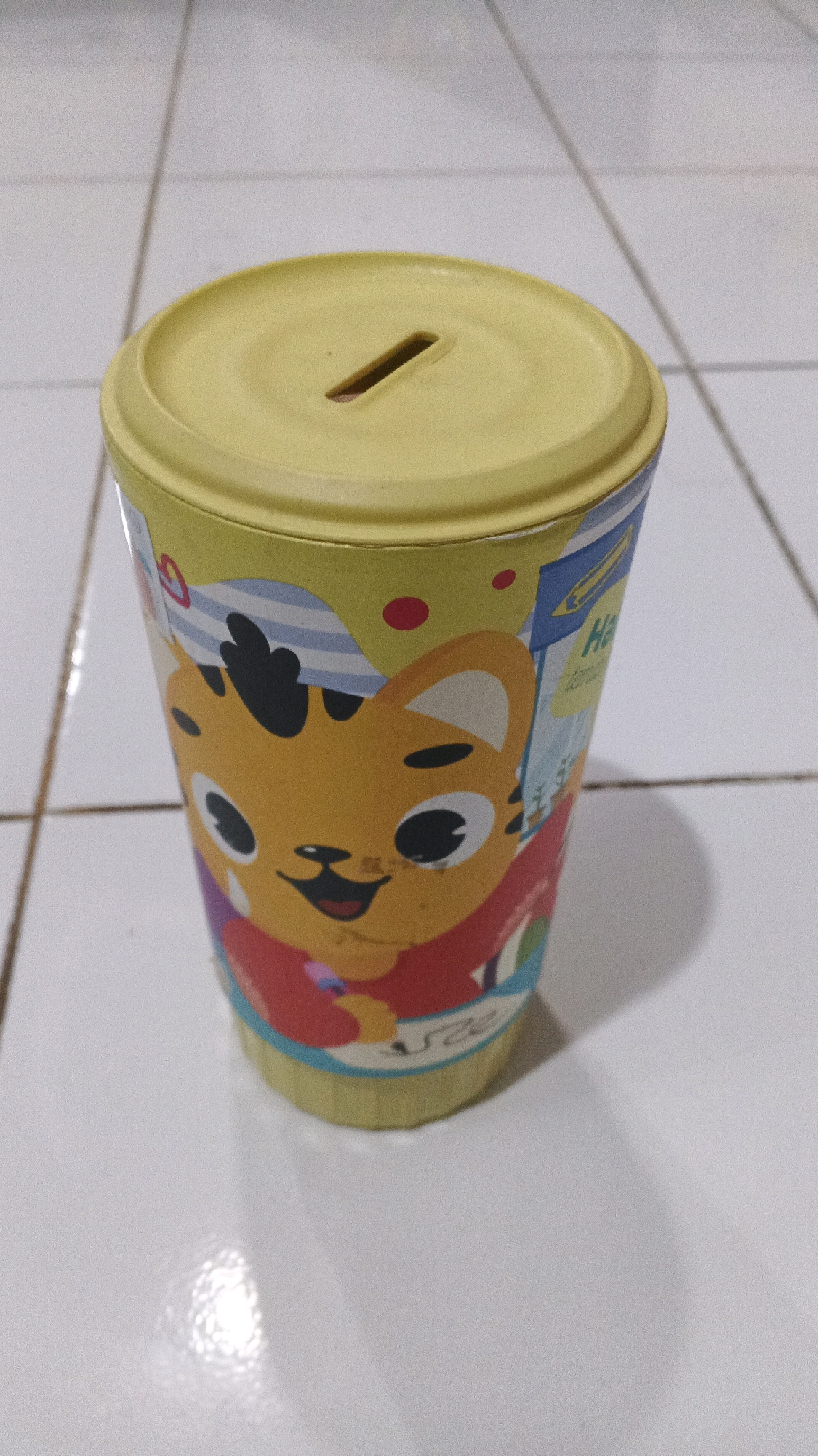 A cylindrical, yellow piggy bank with a cartoon tiger on it sits on a tiled surface in Winong, Indonesia. The piggy bank has a slit in the yellow lid for inserting coins. The tiger on the side of the bank is orange with black stripes and appears to be wearing a red shirt. The background is a light-colored tiled floor with visible grout lines. The lighting is overhead, casting a shadow from the piggy bank onto the floor.