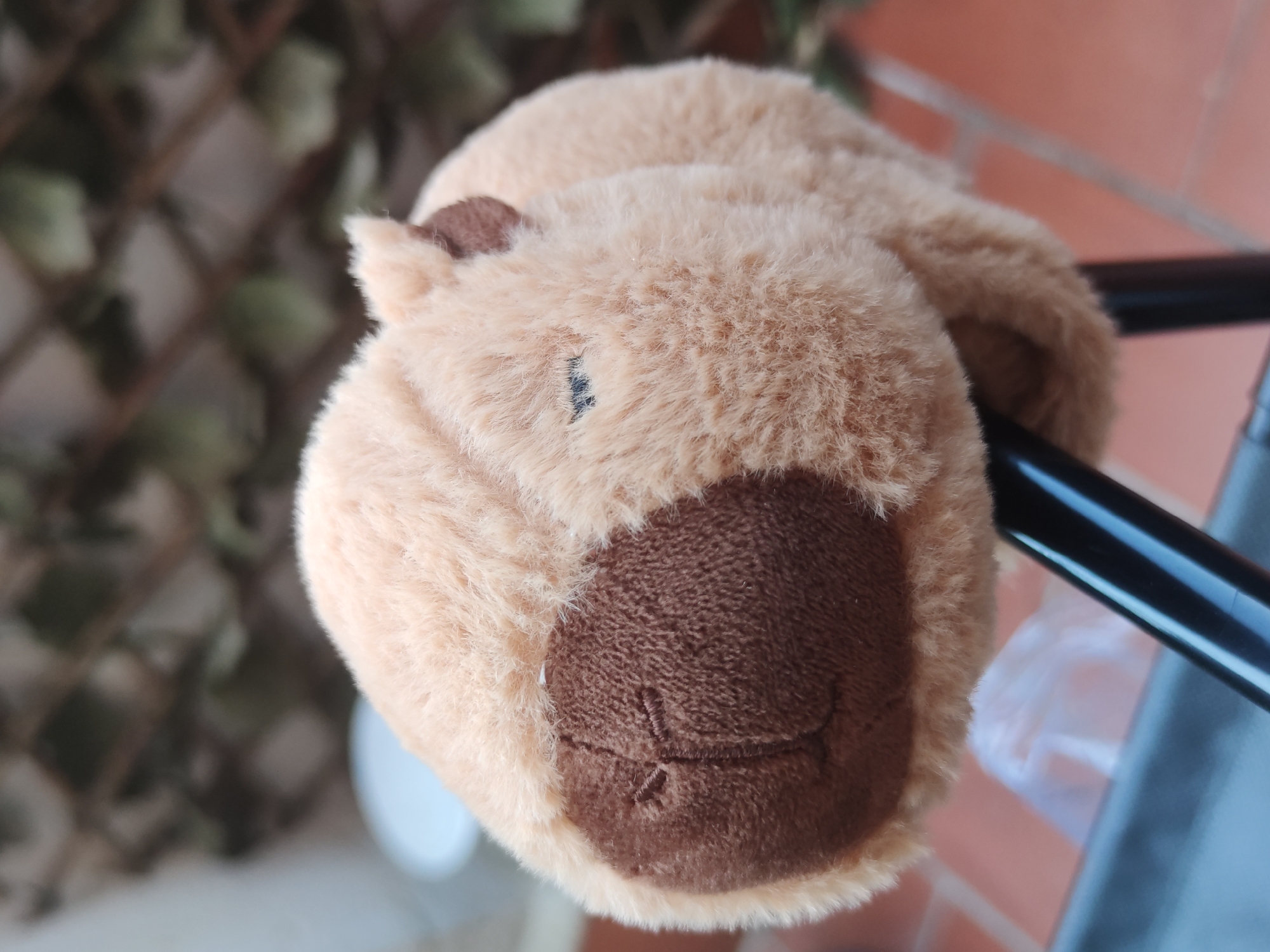 A plush, light-brown capybara toy rests on a black tubular structure, possibly part of a railing or chair. The toy has a darker brown muzzle with stitched details suggesting a smile and a small "x" below the mouth. A single, dark stitched line represents an eye. The background includes an out-of-focus lattice structure with foliage behind it, and a brick wall in the lower right. The scene is likely outdoors. While specific city details are absent, contextual cues suggest it may be within a residential or park area; lack of unique landmarks prevents explicit confirmation as Barcelona.