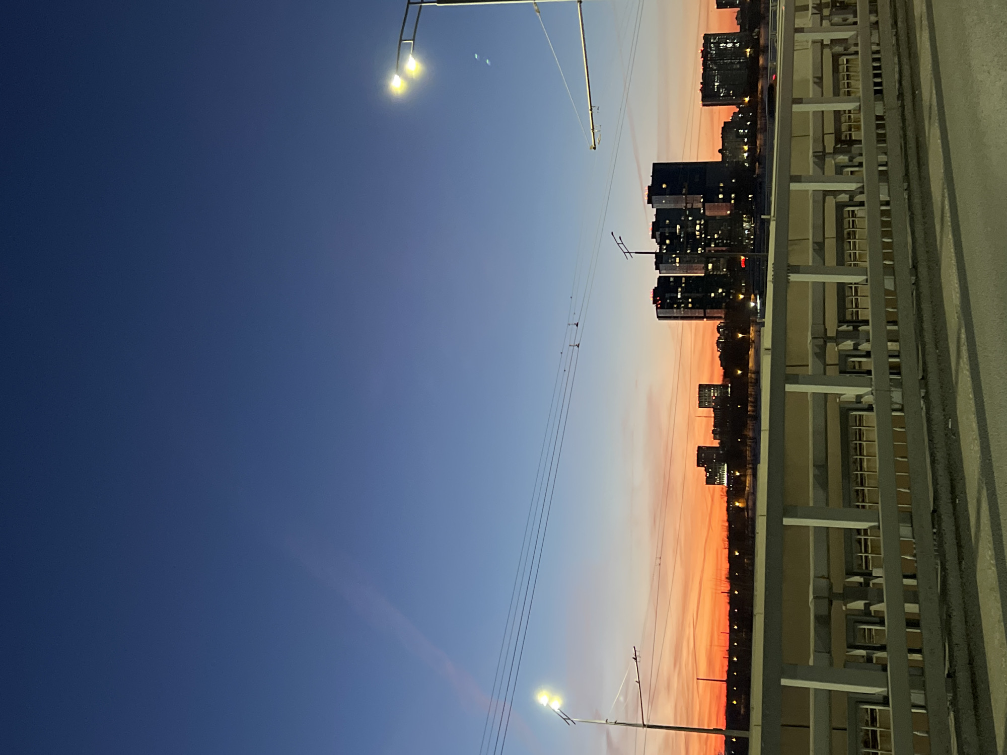 The image depicts a twilight scene over the city of Kazan, Russia, viewed from a bridge. The sky transitions from a deep blue at the zenith to a vibrant orange and red at the horizon. Faint linear streaks are visible in the upper blue sky.
In the foreground, a dark grey asphalt road surface occupies the bottom portion of the frame. Above it, a light grey concrete barrier or railing structure with vertical posts and horizontal rails extends across the image. Beneath this railing, a sub-structure with multiple rectangular openings, possibly windows or structural elements, is visible.
The midground features a city skyline silhouetted against the colorful horizon. A prominent cluster of three dark, multi-story buildings with numerous illuminated windows stands slightly left of center. Additional buildings with illuminated windows are visible to the right. A line of scattered lights marks the distant horizon. This cityscape is consistent with views of modern developments in Kazan, specifically the Kazan Riviera area, likely viewed from a bridge crossing the Kazanka River. Distant ground appears dark with sparse, leafless vegetation and possible snow cover.
Artificial lighting includes two streetlights with dual illuminated yellow/white lamps on tall poles, one on the far left and one on the far right. Multiple overhead utility lines, possibly power or tram lines, traverse horizontally across the mid-sky.
No individuals are visible. A faint red streak on the road surface in the midground on the right indicates the movement of a vehicle's taillights. The overall lighting suggests the time of day is twilight, likely after sunset.