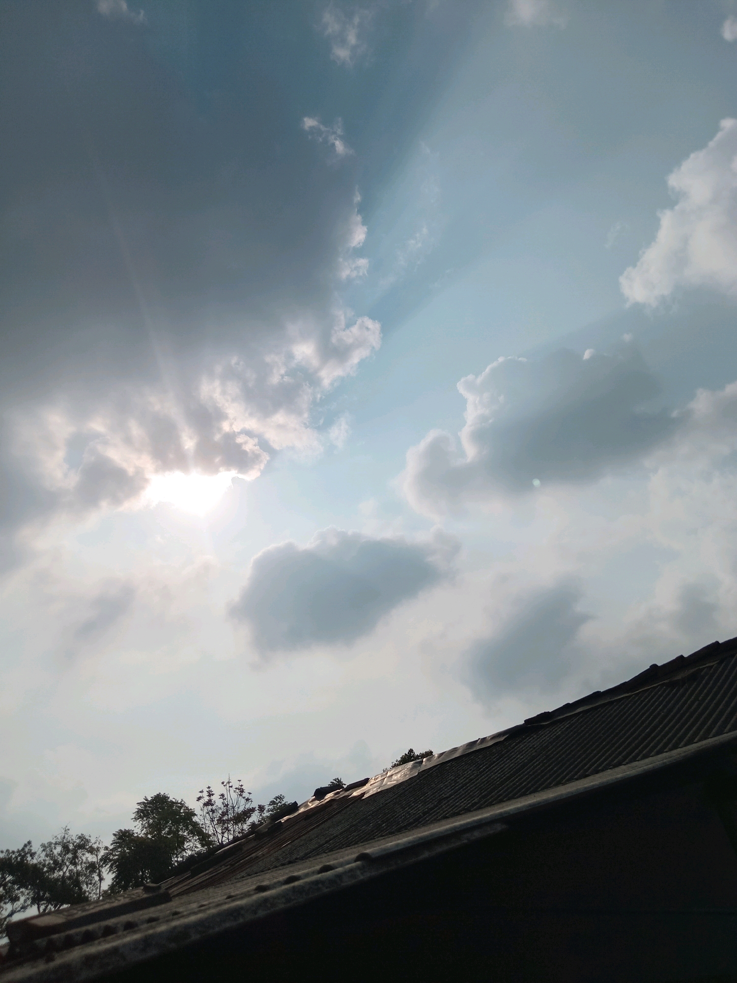 The image, taken in Claket, Indonesia, depicts a cloudy sky with visible sunbeams breaking through. The sun is partially obscured by clouds, creating a bright, luminous area in the upper left quadrant. The sky transitions from darker, denser clouds on the left to lighter, more diffused clouds towards the right, with hints of blue peeking through. In the foreground, a dark, textured roofline dominates the lower portion of the frame, with the corrugated material and some foliage visible. The overall scene suggests an outdoor environment, likely a residential area, given the presence of the roof. There are no people or animals visible in the image.